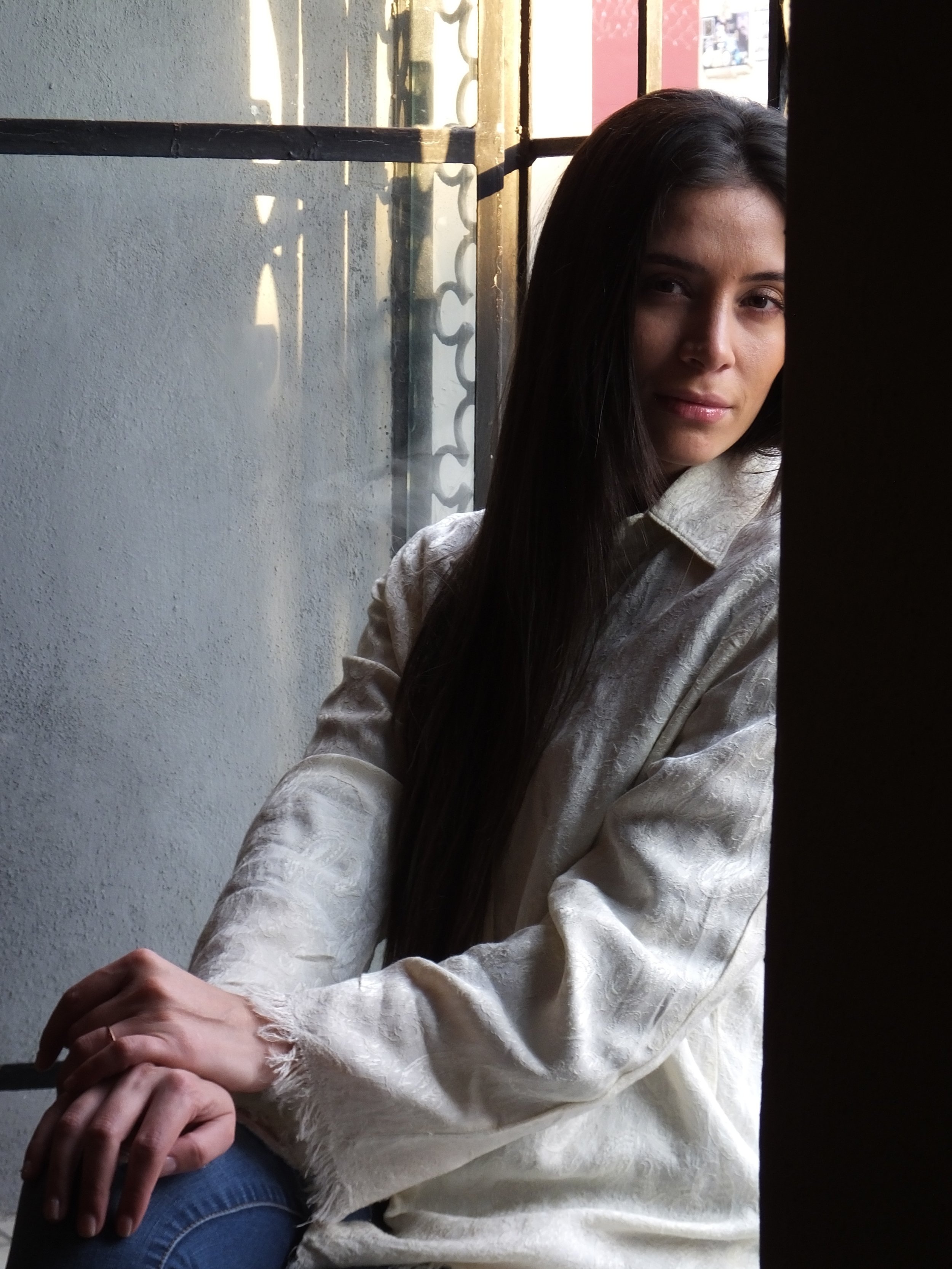 A young woman with long dark hair sitting by a window, looking at the camera, with a light-colored blouse and jeans.