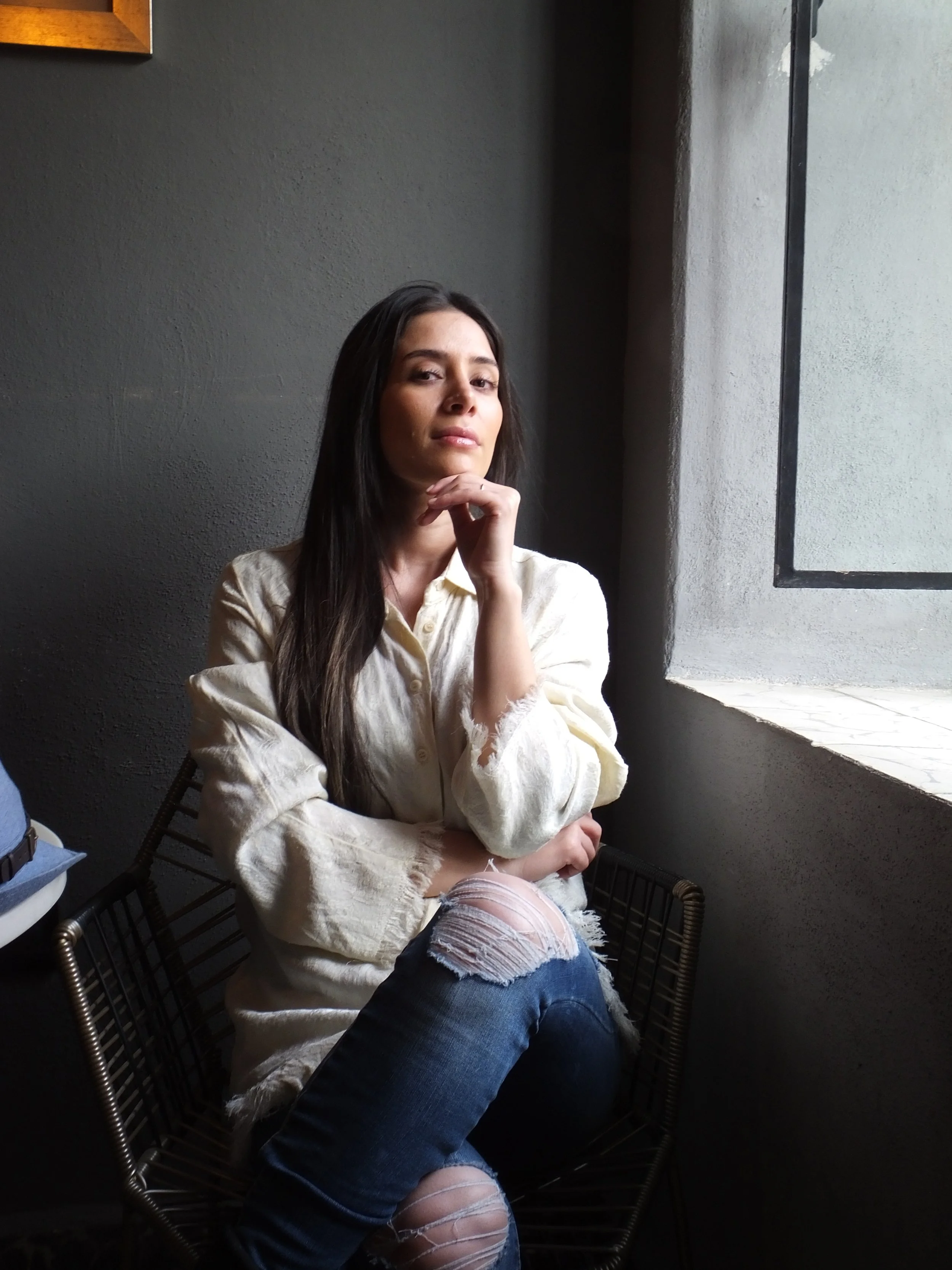 A woman with long dark hair sitting on a wireframe chair indoors near a window, wearing a light-colored shirt and ripped jeans, with her left hand resting on her chin and looking at the camera.