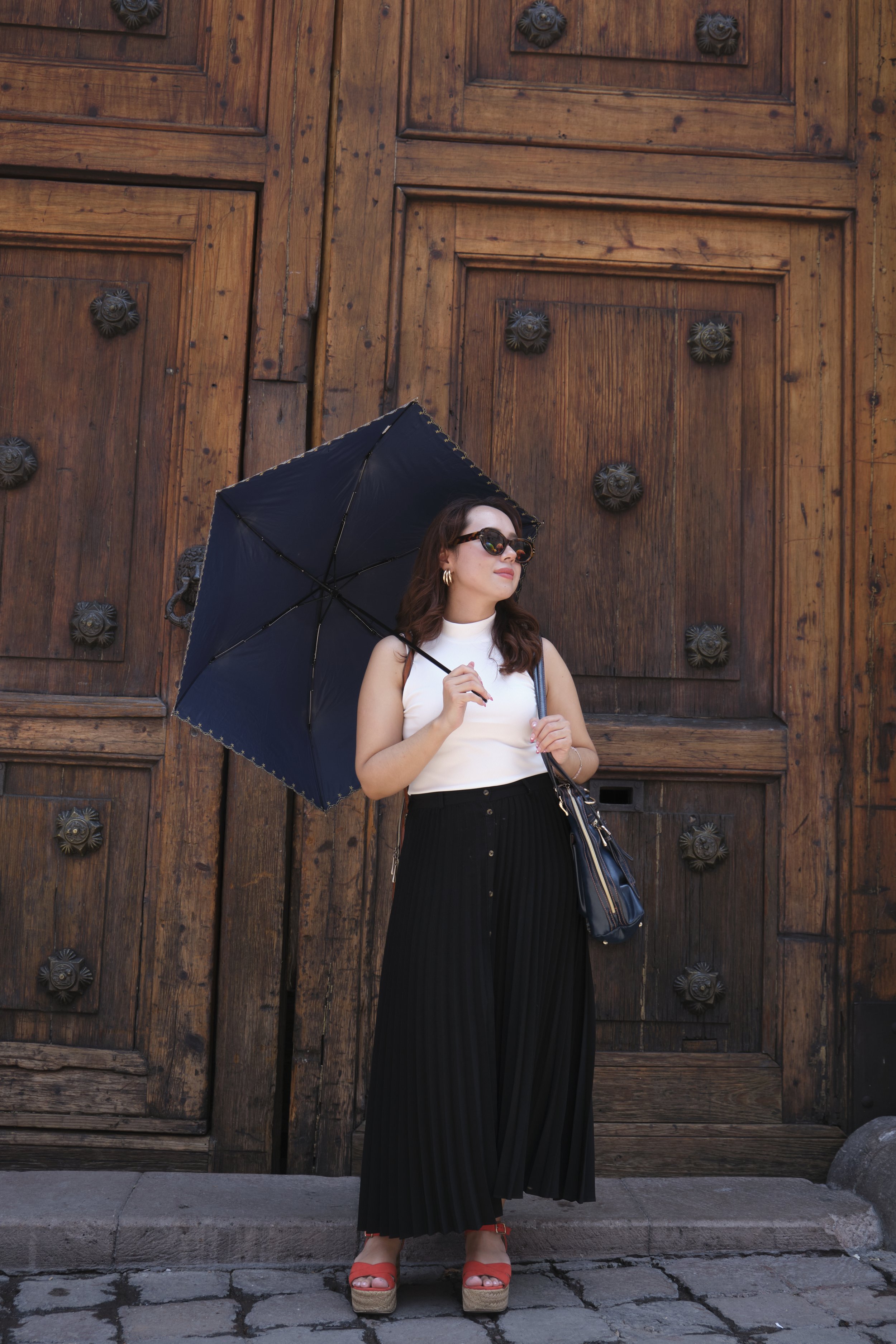 A woman standing in front of a large wooden door, holding a black umbrella, wearing sunglasses, a white sleeveless top, black pleated wide-leg pants, and red wedge sandals, with a black handbag.