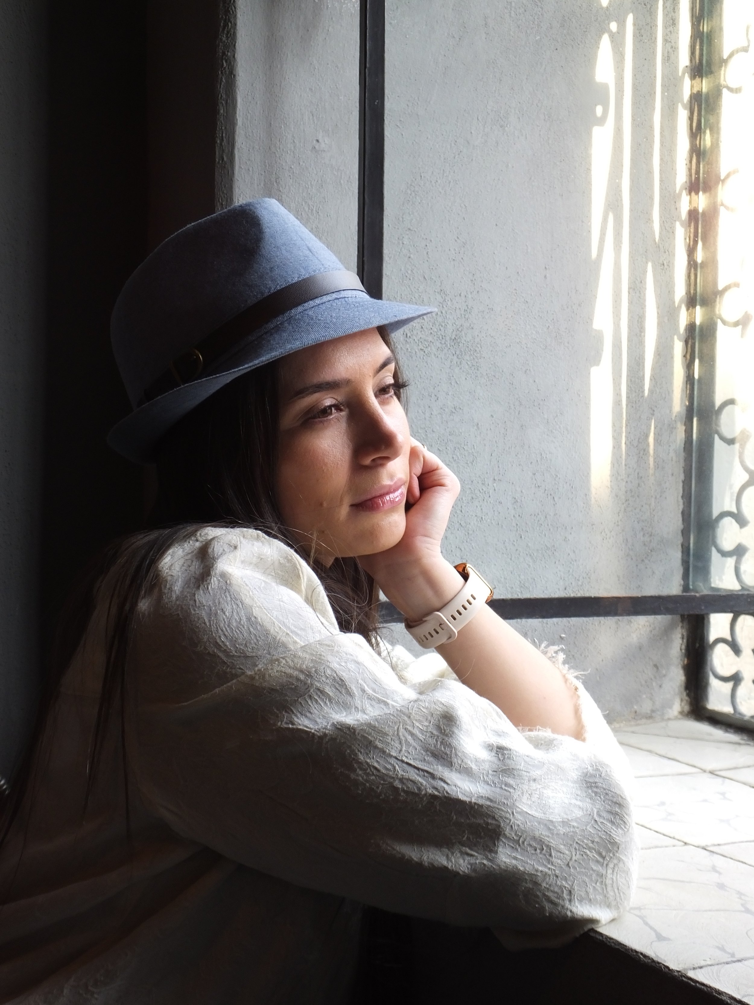 A woman wearing a gray hat with a dark band and a white long-sleeve shirt, sitting by a window with sunlight streaming in, looking contemplative with her chin resting on her hand.