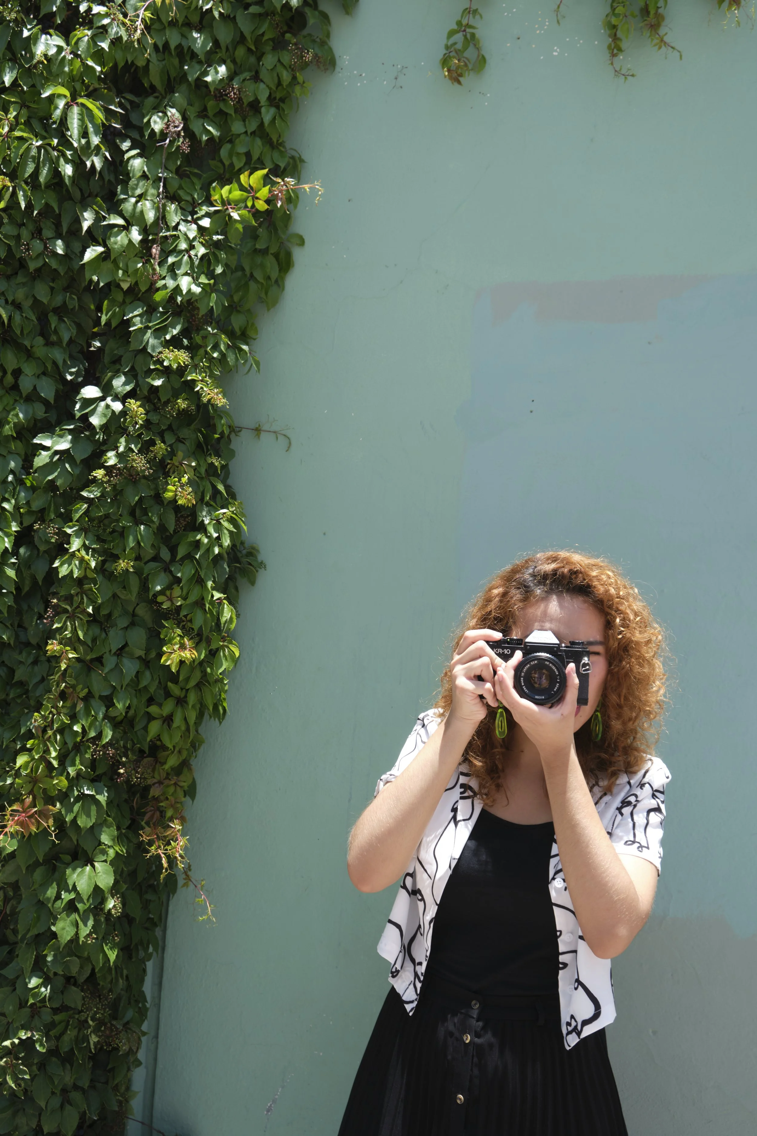 A woman with curly hair takes a photo with a camera in front of a green wall and ivy-covered corner.
