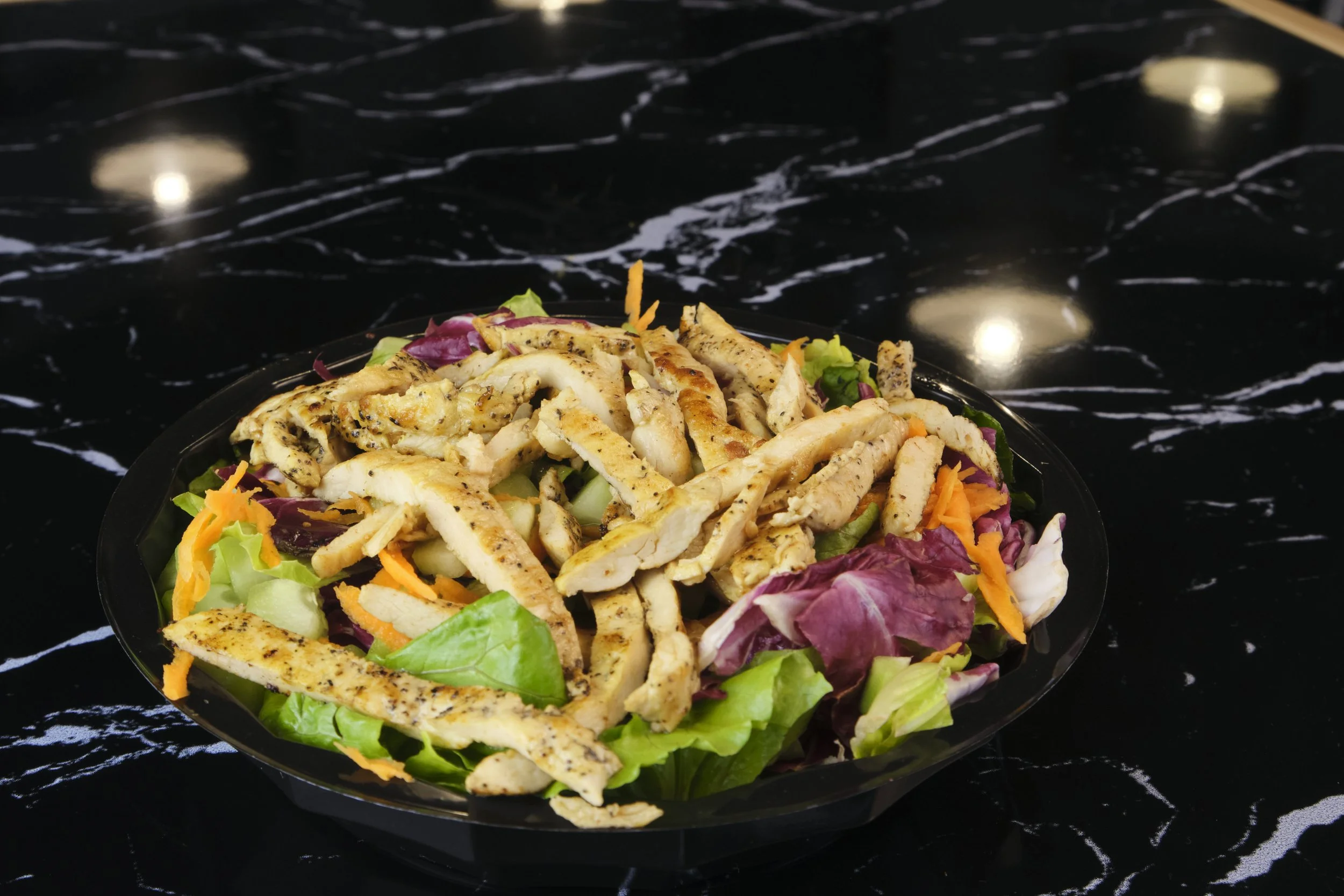 A chicken Caesar salad with grilled chicken strips, lettuce, shredded carrots, purple cabbage, and other mixed greens in a black bowl on a black marble countertop.