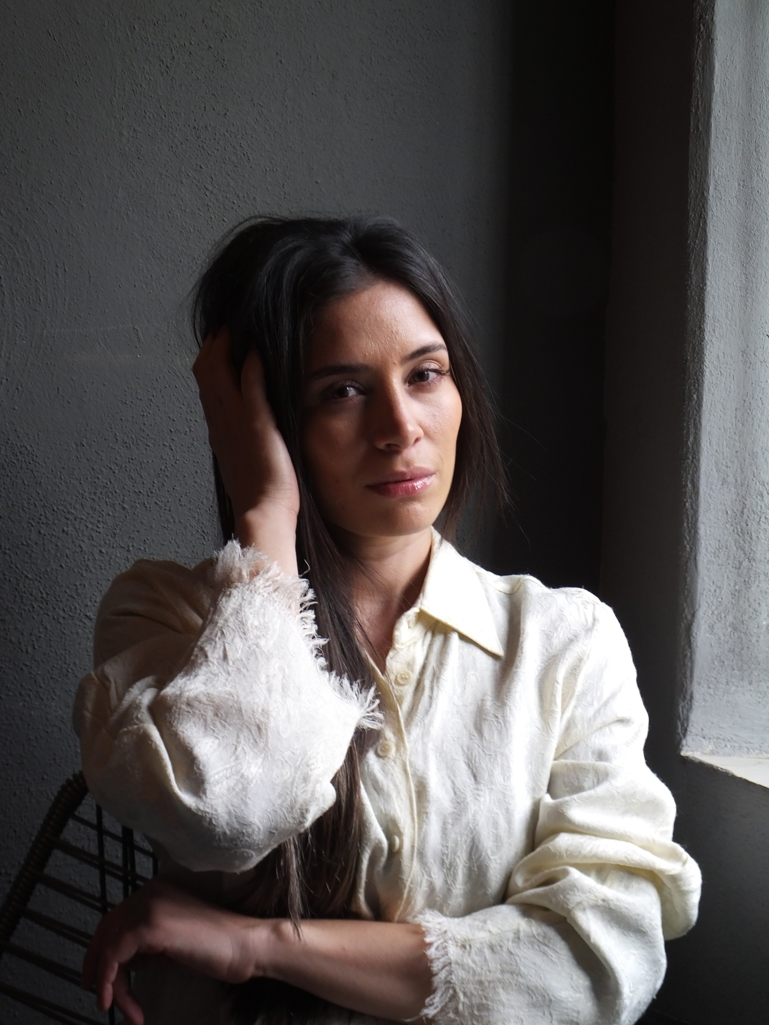 A woman with long dark hair sitting near a window, touching her head with her right hand, wearing a cream-colored fluffy sweater, looking at the camera with a neutral expression, in a dimly lit room.