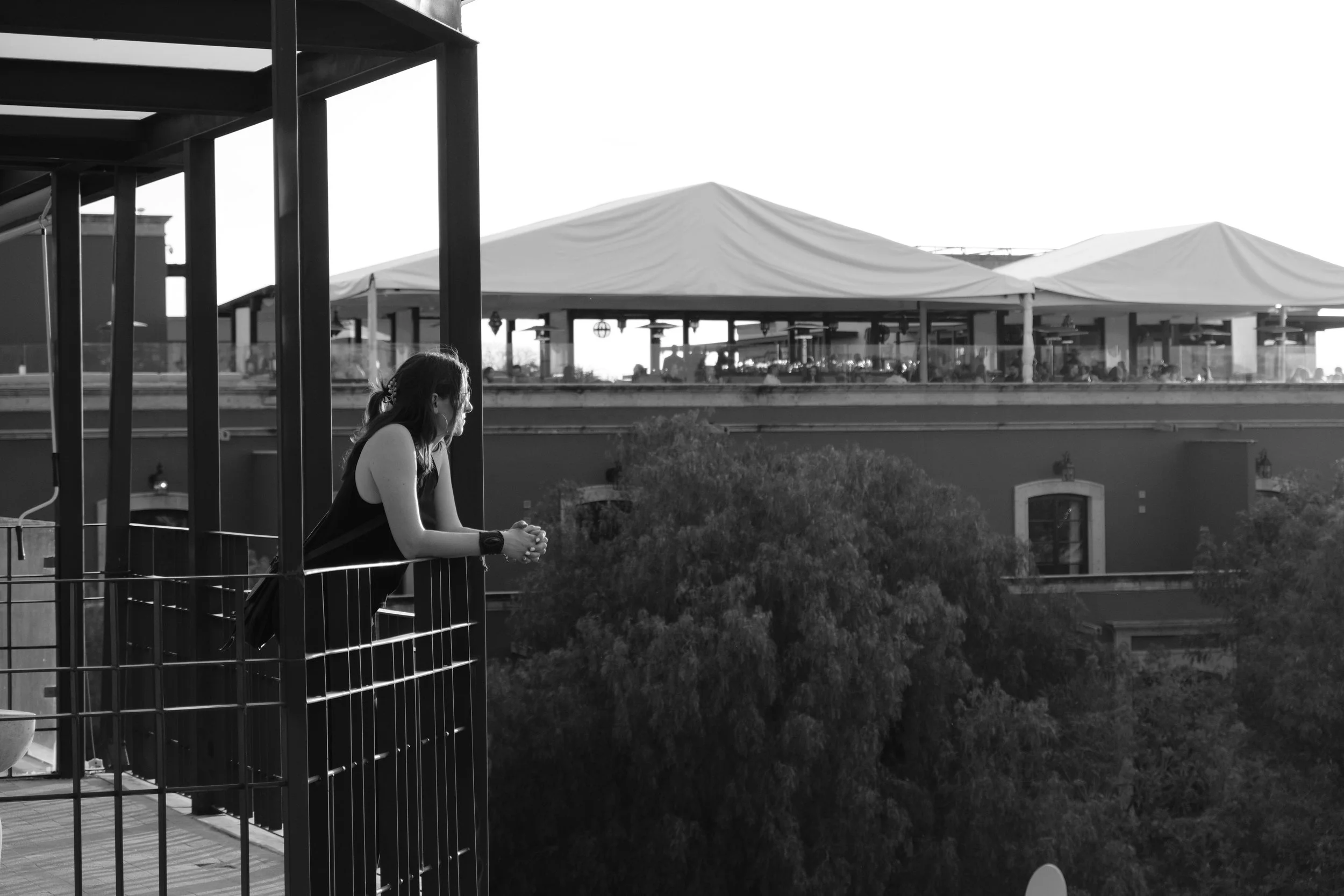 A woman with a ponytail and tank top leaning on the railing of a balcony pondering, looking out toward a Rooftop dining area with tents and people in the background.