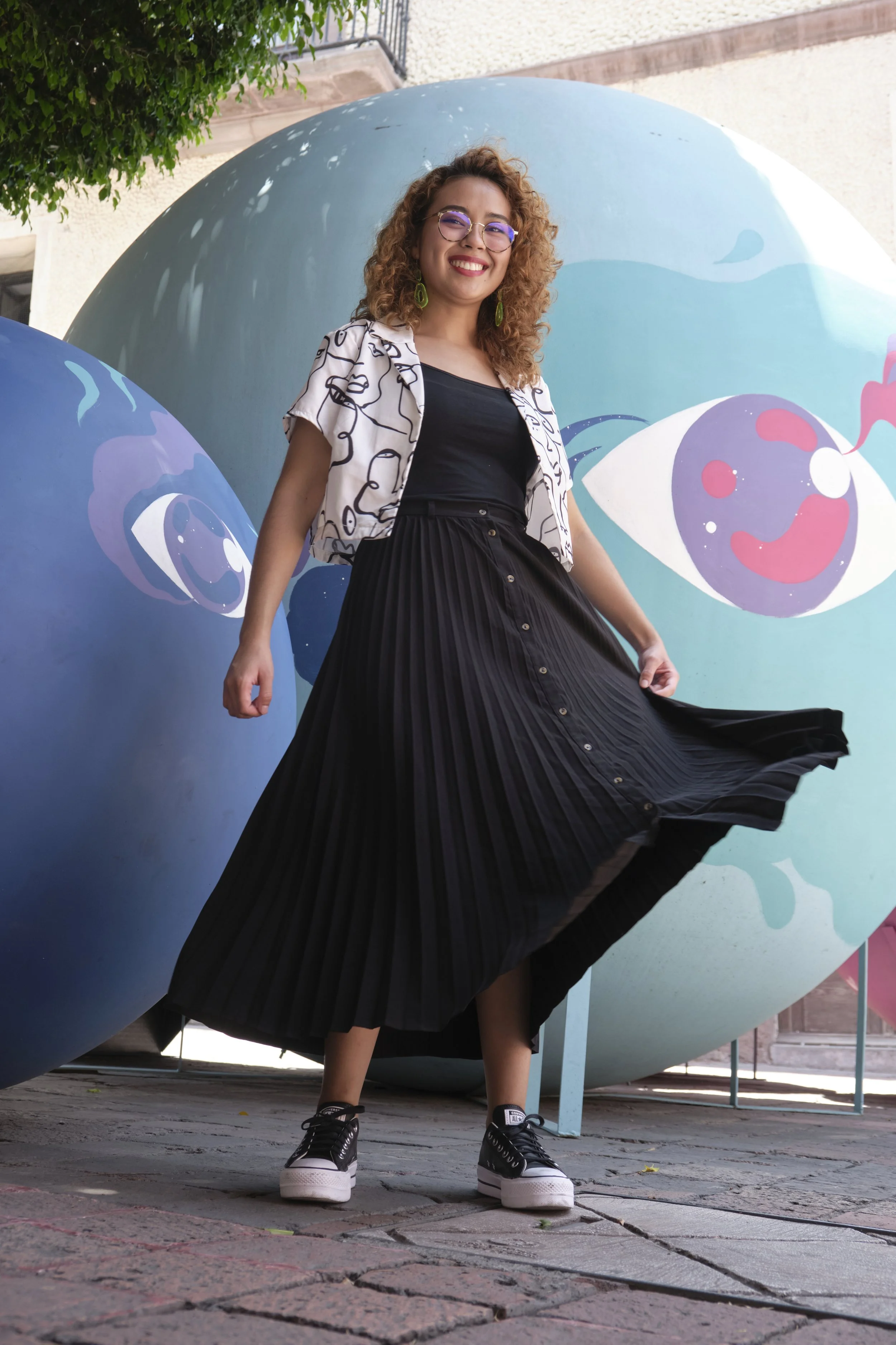 A woman with curly hair, glasses, and earrings, smiling and posing in front of large colorful spherical art installations with eye designs.