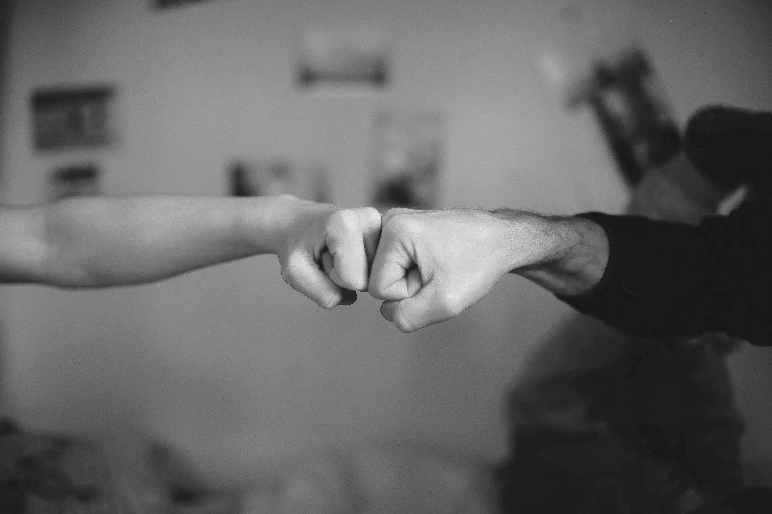 Black and white photo of two hands fist-bumping, one with a lighter skin tone on the left and one with a darker skin tone on the right, in a room with blurred background.