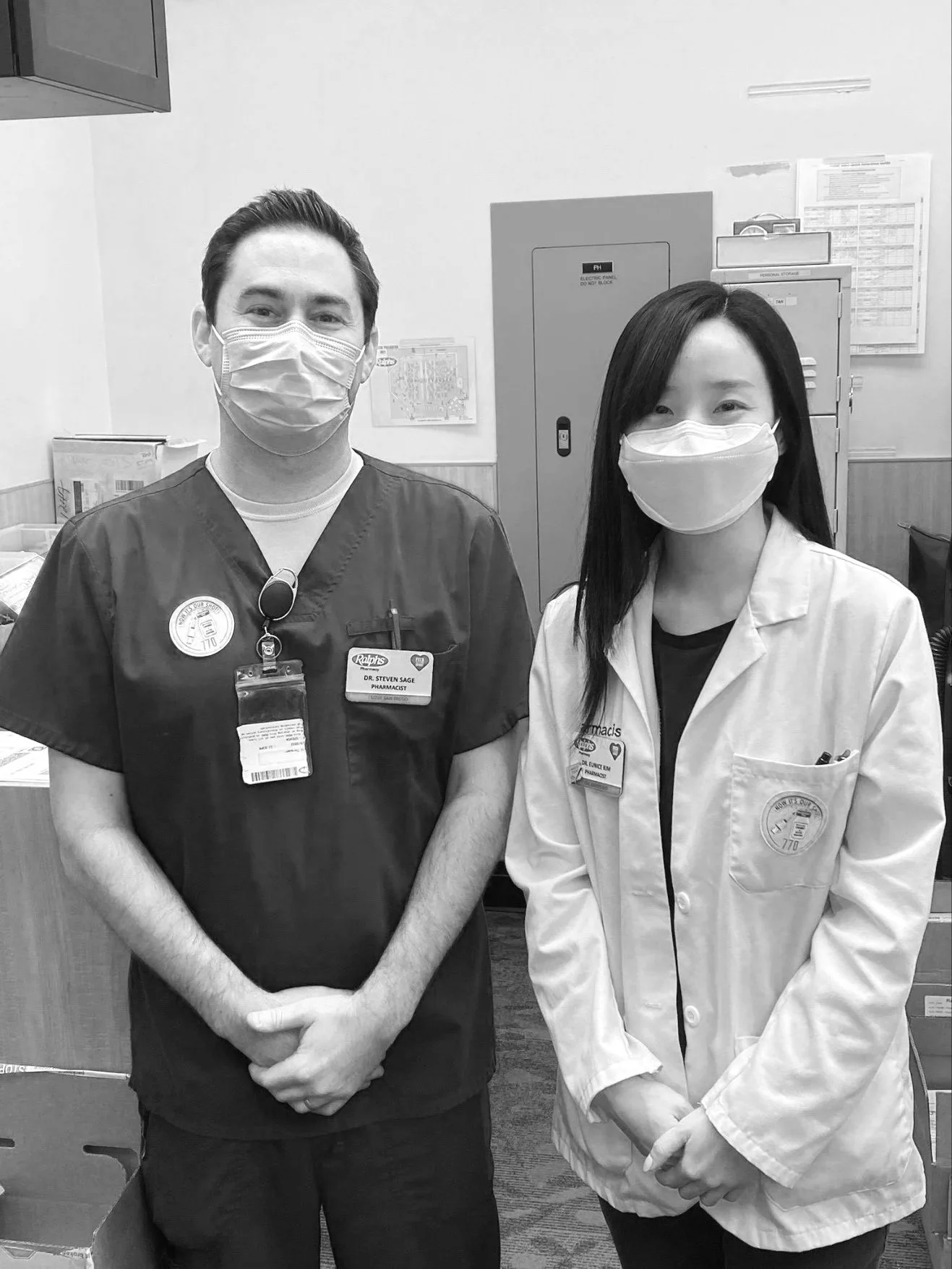Two healthcare professionals, a male and a female, pose together in a medical setting. They are both wearing masks, scrubs, and lab coats.