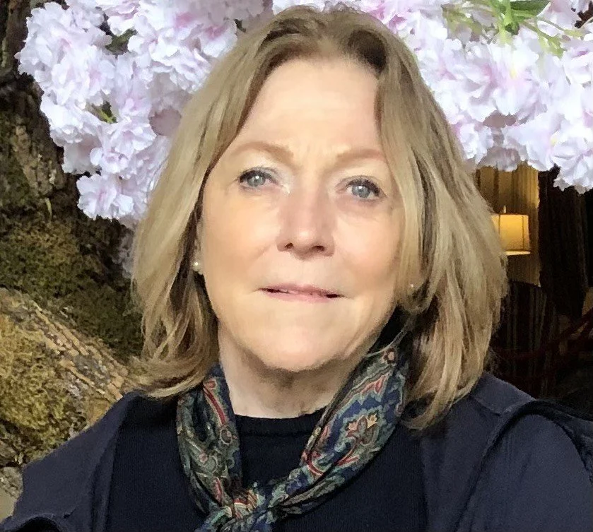 A woman with shoulder-length light brown hair and blue eyes standing in front of a background of pink hydrangea flowers and a tree trunk.