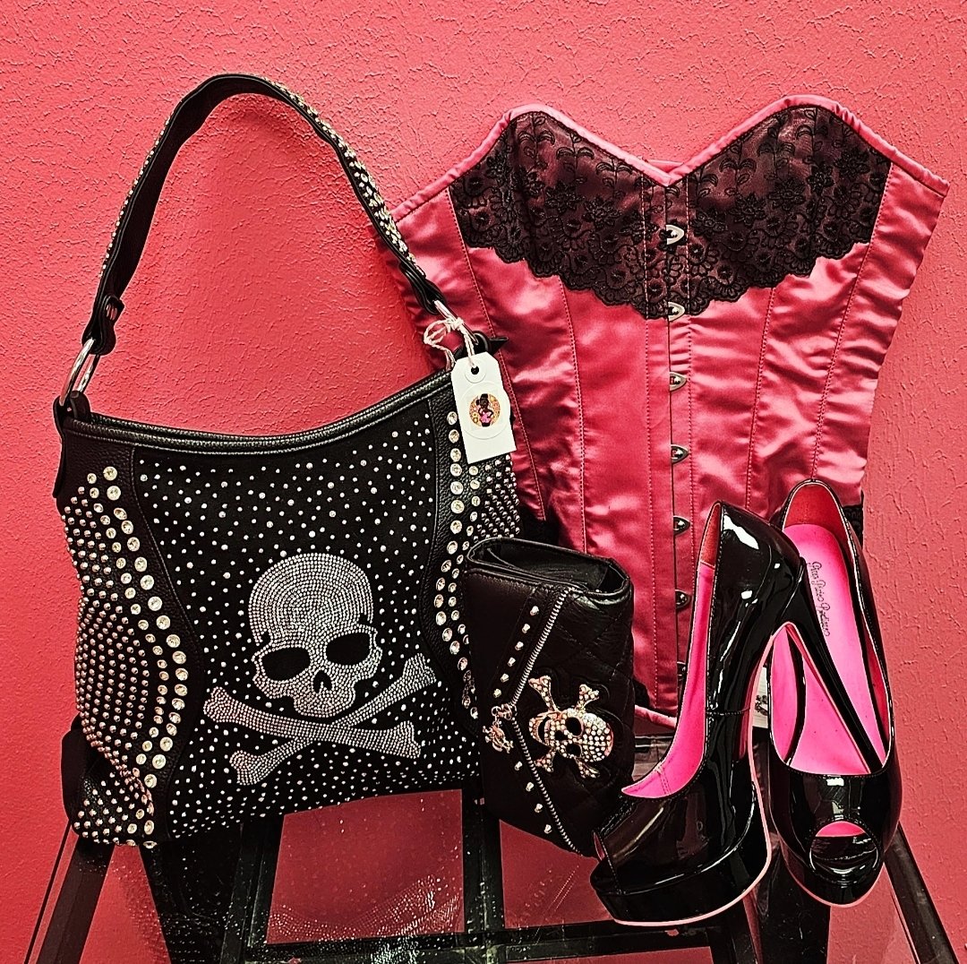 A pink corset with black lace detailing, a black handbag and wallet with skull and crossbones designs, a pair of black and pink high-heeled shoes, all arranged against a pink textured wall.
