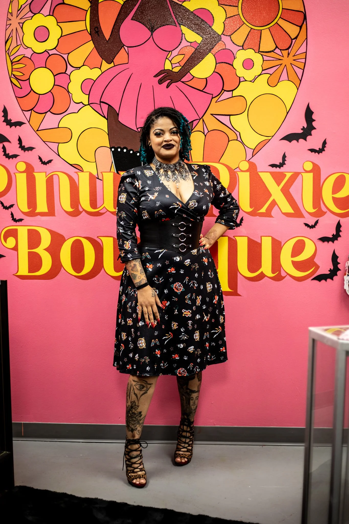 A woman with tattoos wearing a black dress with a floral pattern and lace-up high heels posing in front of a colorful pink wall with a large cartoonish image of a woman holding a pink umbrella surrounded by yellow and orange flowers and flying bats.