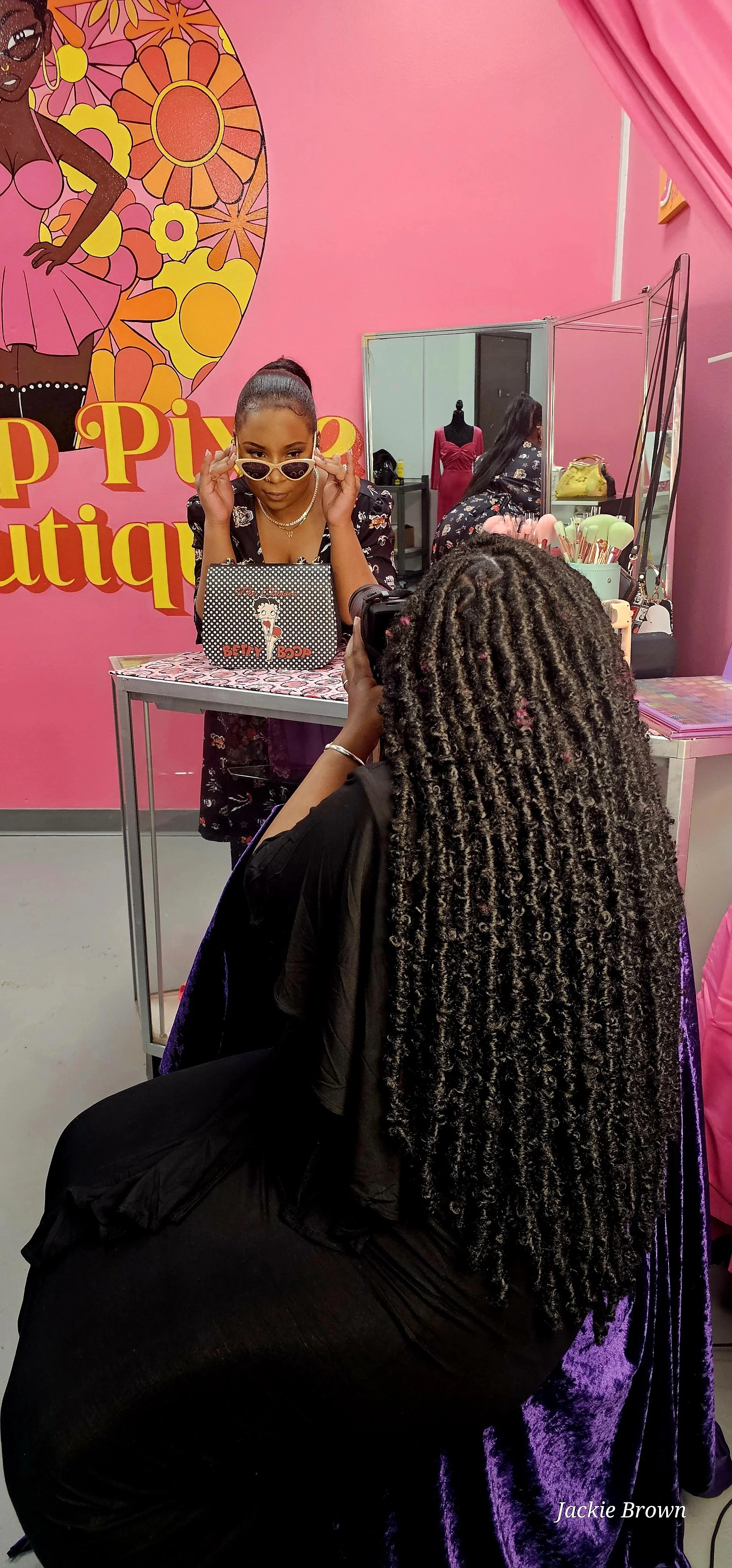 A woman with long curly hair is sitting at a table recording a woman posing for a photo in front of a bright pink wall with a large artistic mural of a woman in a pink dress and colorful flowers. The woman posing is leaning on a table with a Betty Boop-themed bag, wearing sunglasses and a black floral outfit. There are makeup items and a mirror on the table. The setting appears to be a boutique or photobooth area.