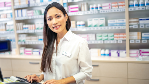 Smiling pharmacist at the counter with shelves of medicine in the background.