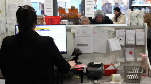Pharmacy staff member using a computer behind the counter while patients wait at the pharmacy reception.