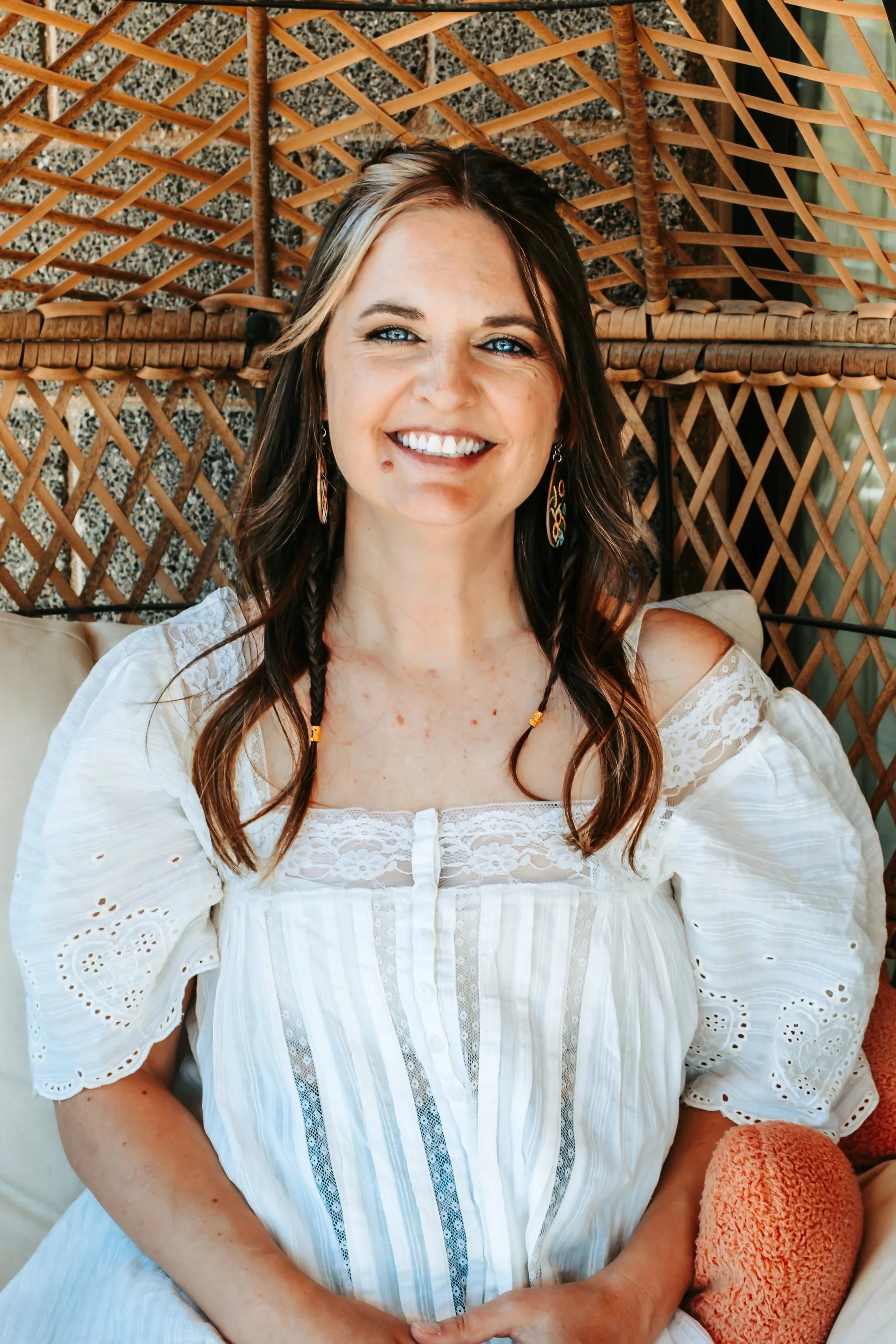 A woman with brown hair and blue eyes smiling, wearing a white lace dress with cut-out details, sitting on a cushioned seat with a wicker or rattan backdrop.