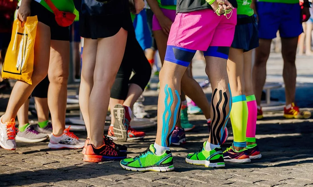 Group of runners standing before a race with kinesiology tape applied to lower legs to support shin splint prevention.