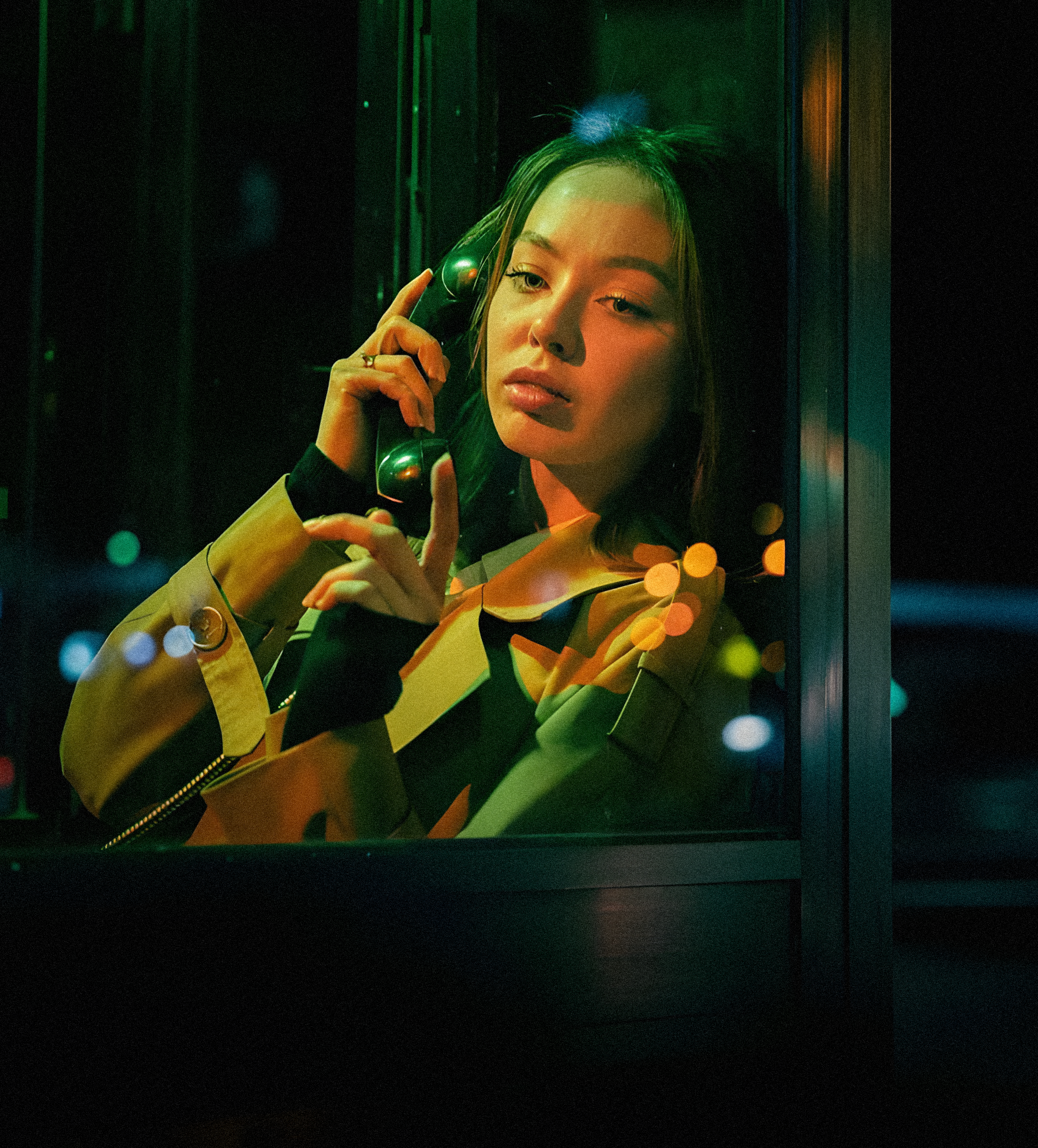 A young woman with long dark hair and makeup talking on a black vintage telephone, framed by a window with colorful bokeh lights at night.