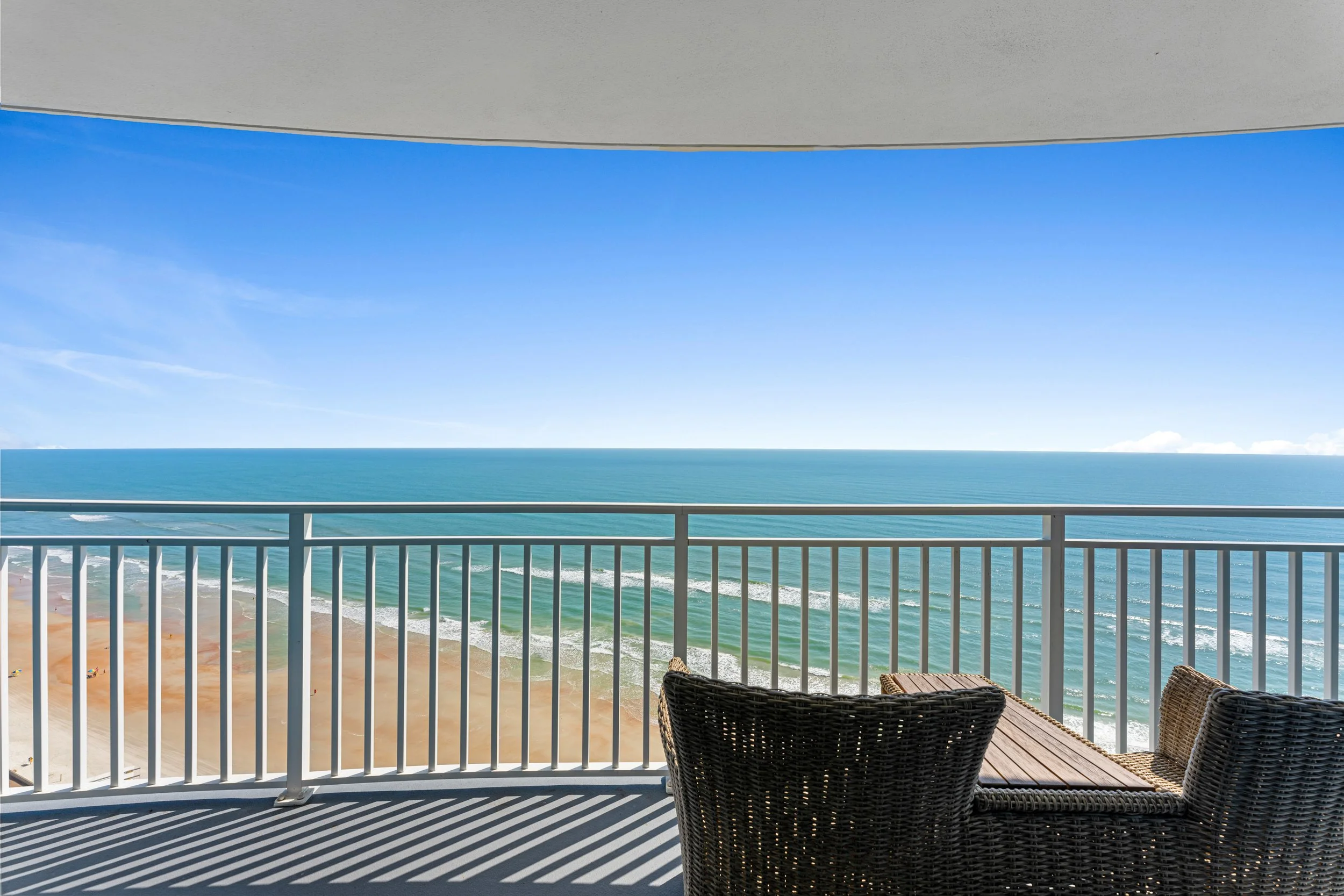 Balcony overlooking a sandy beach, ocean waves, and a clear blue sky with a few clouds.