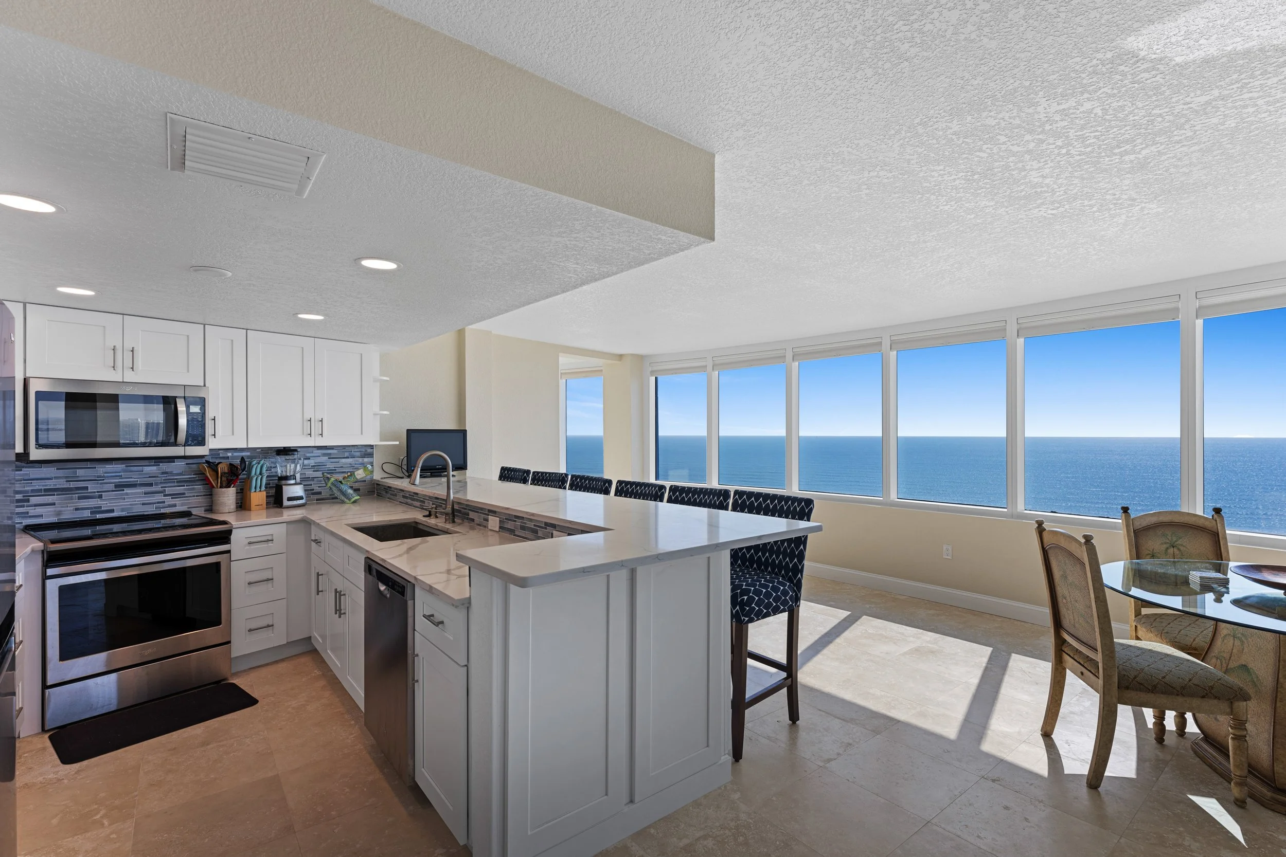 Kitchen with white cabinets, stainless steel appliances, and a bar area with four black patterned chairs. The space opens to a dining area with a glass-top table and patterned chairs. Large windows overlook the ocean with a clear blue sky.
