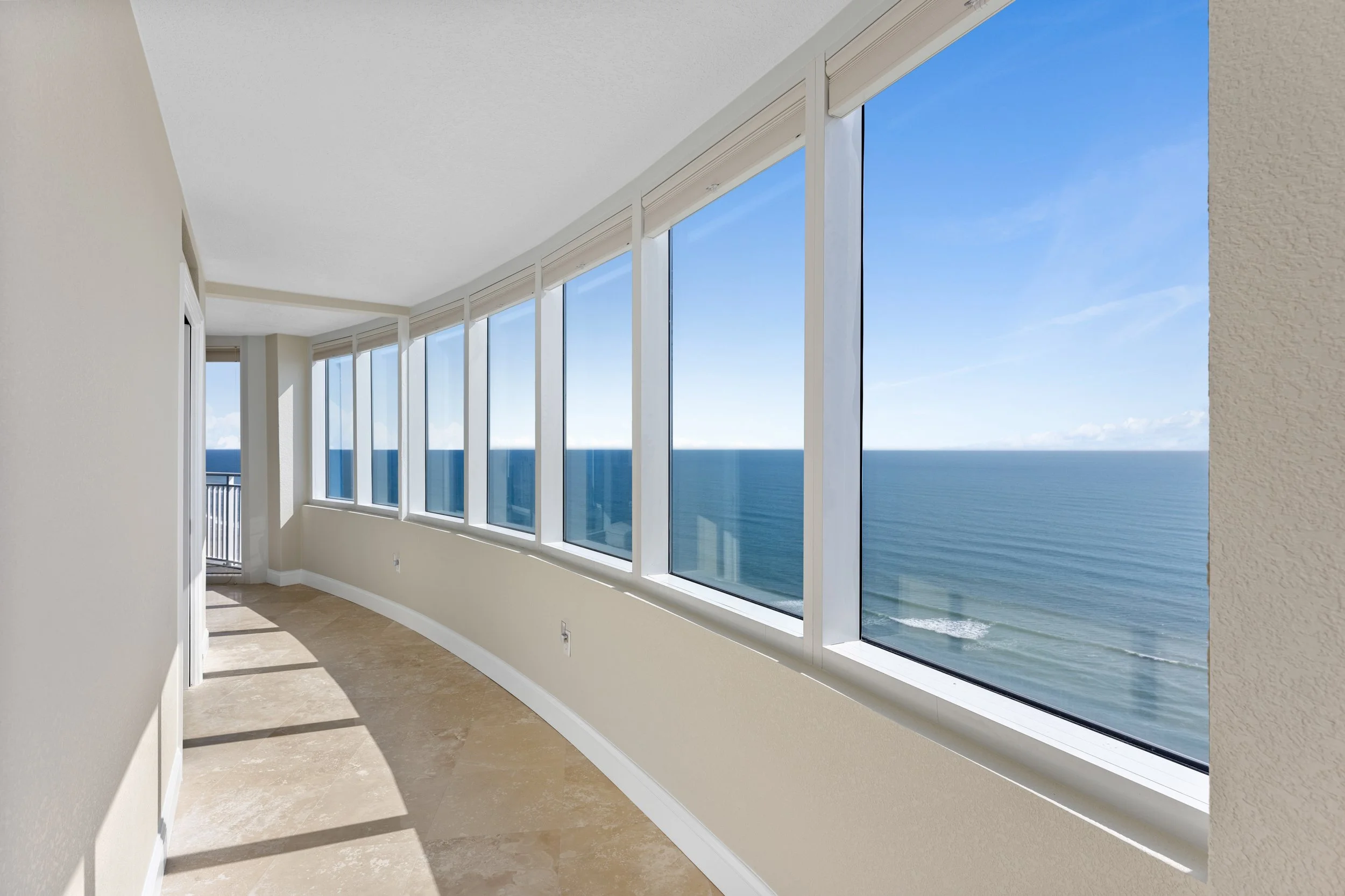Empty corner room with large windows overlooking the ocean, beige walls, and tiled floor.