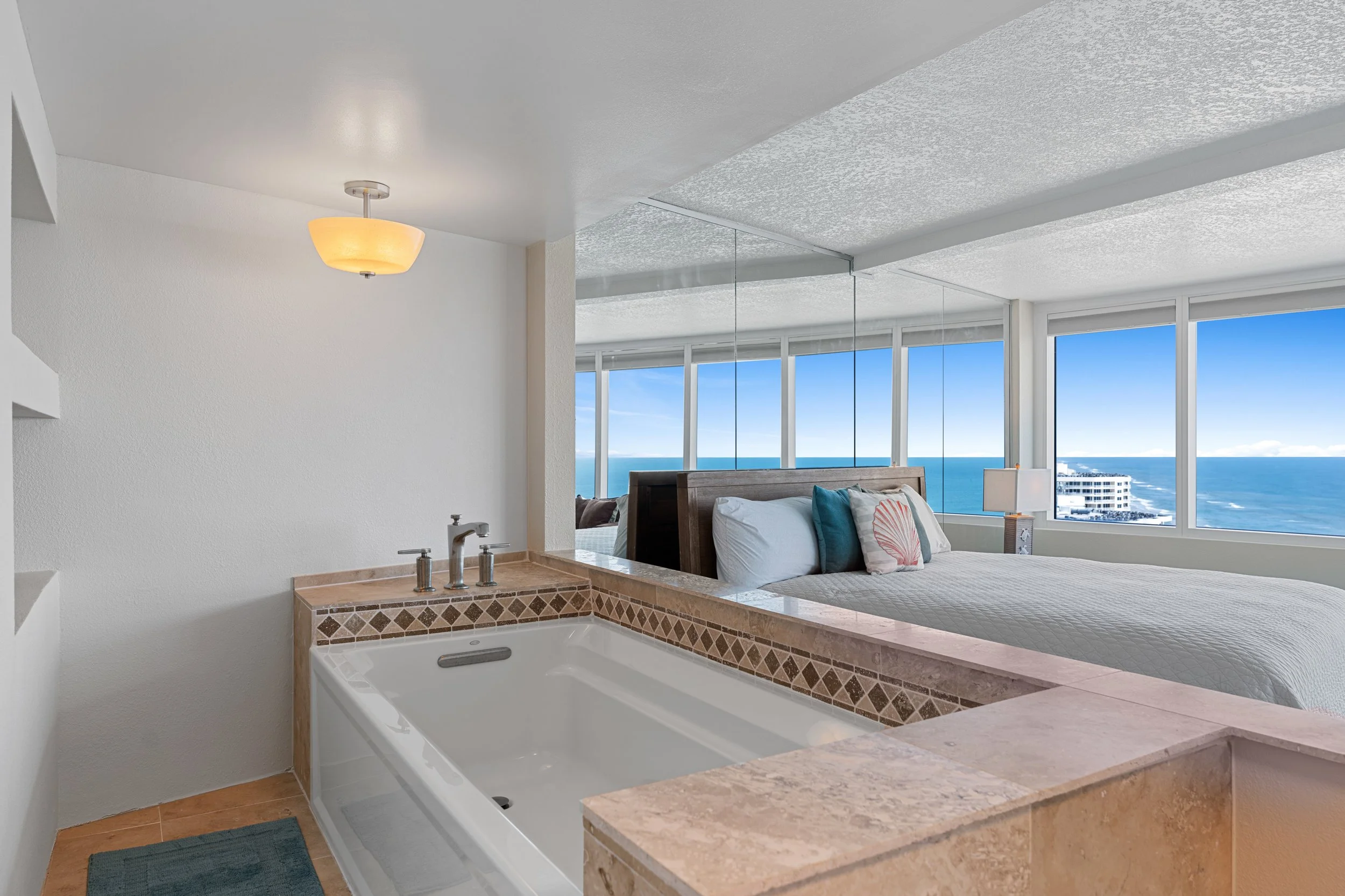 Room with a large window view of the ocean, a bed with pillows, a bedside lamp, and a bathtub with decorative tile.