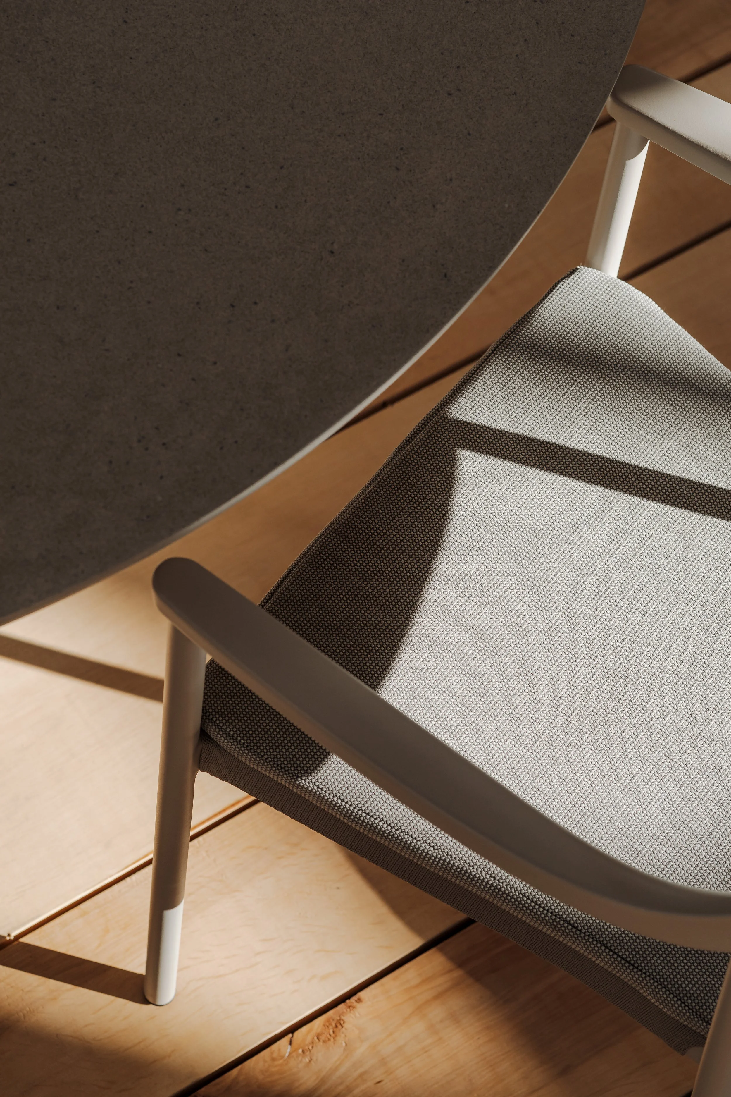 Close-up of a modern chair with a mesh seat and white frame, next to a gray table on a light wooden floor with sunlight creating shadows.