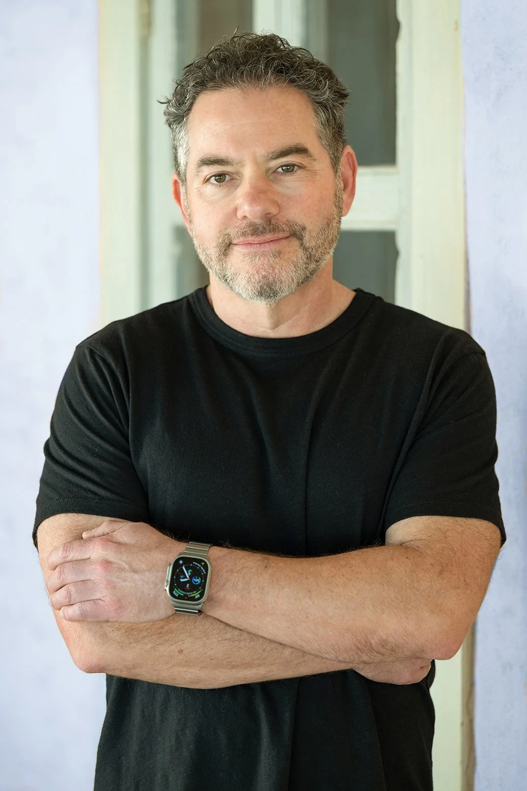 A middle-aged man with gray hair and beard, wearing a black t-shirt and smartwatch, standing outdoors with arms crossed, smiling gently at the camera.