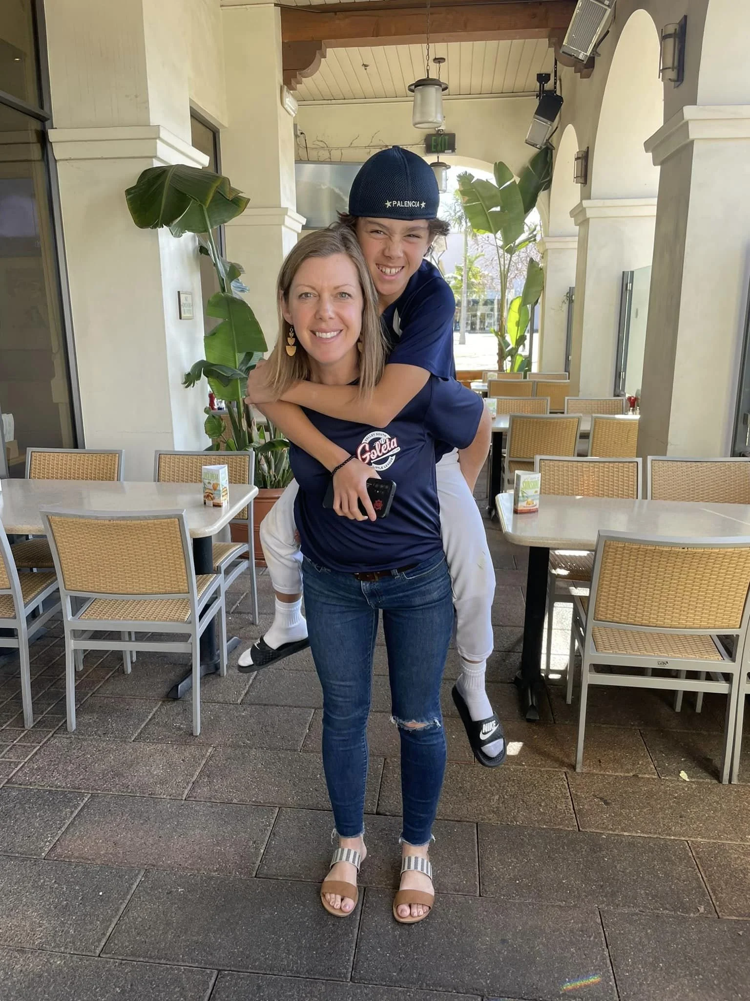 A woman with shoulder-length hair wearing jeans and sandals is standing and smiling, with a young boy in a blue shirt and white pants on her back, giving a piggyback ride, making a funny face. They are inside a restaurant or cafe with outdoor seating and large potted plants.
