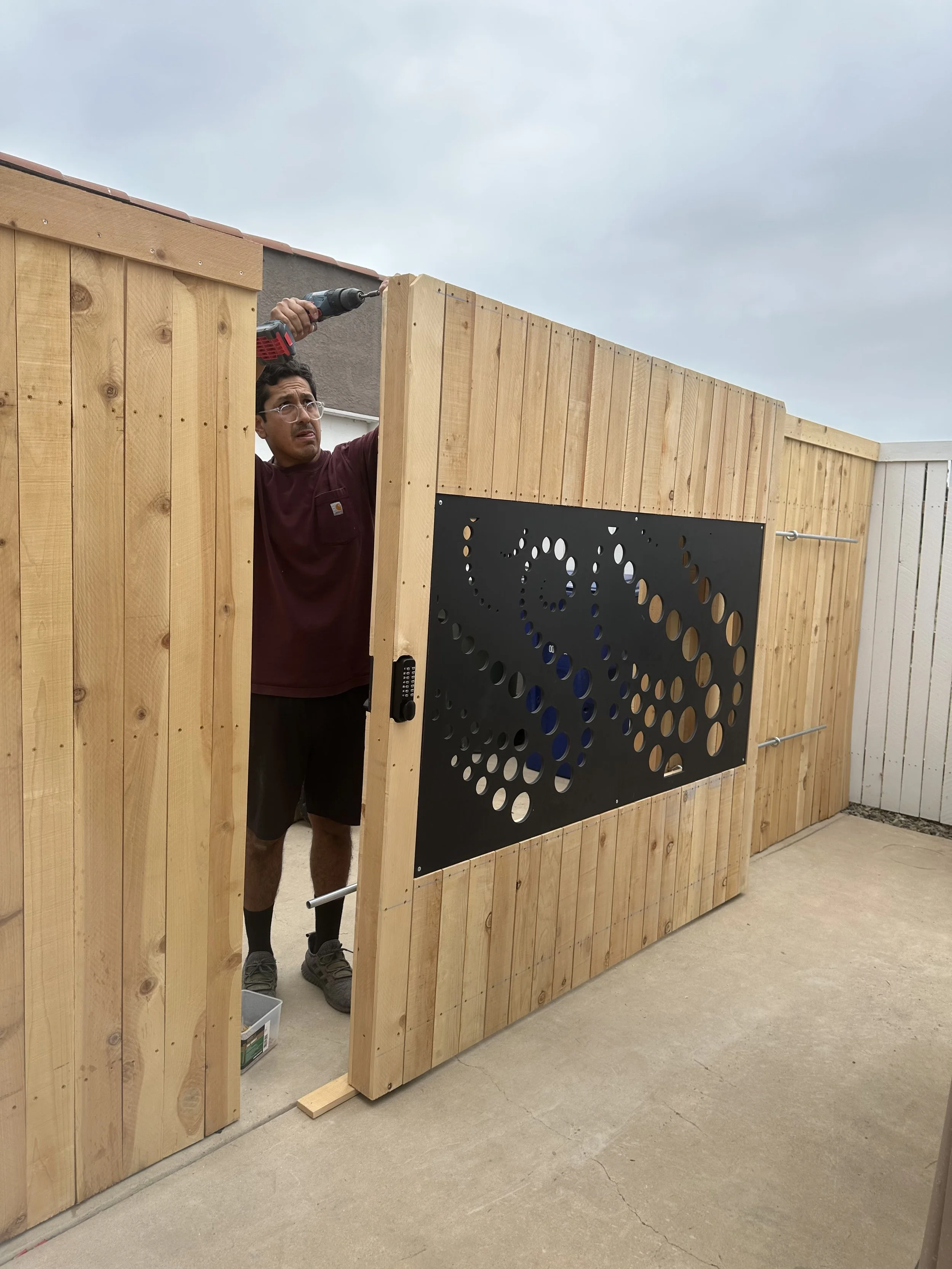 A person working on a wooden fence with a power drill, partially seen through an open gate.