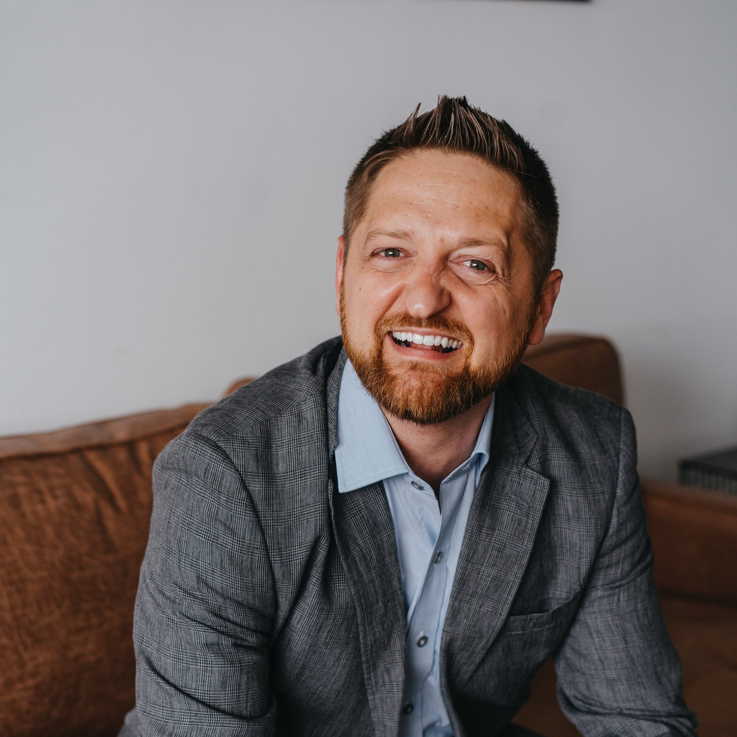 A middle-aged man with a beard and short hair smiling and wearing a suit, sitting on a brown couch in a room with a plain light-colored wall.