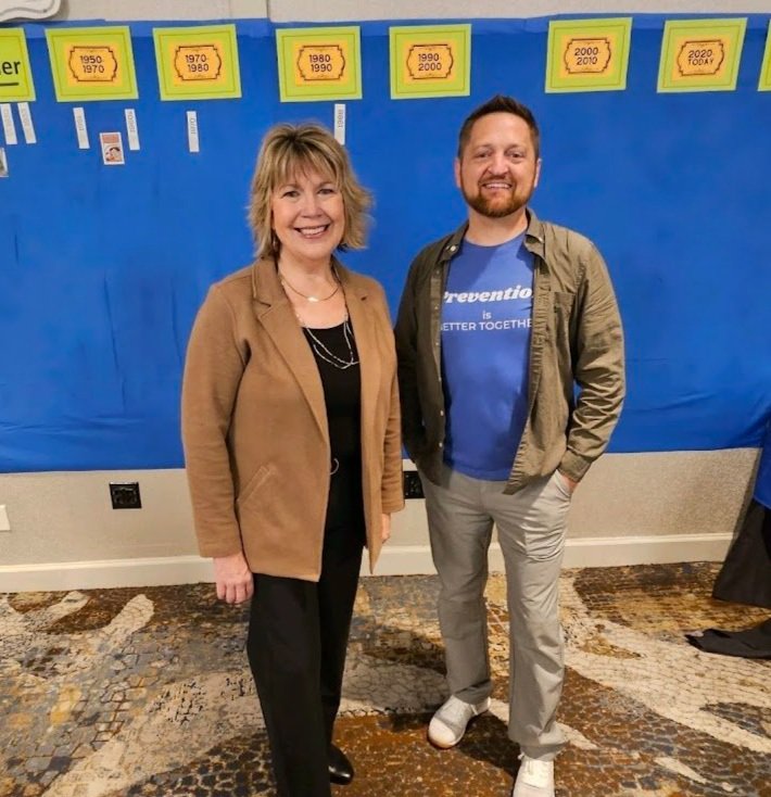 Two smiling women standing together in a room with a blue wall decorated with yellow and white timeline banners. One woman has short blonde hair and is wearing a brown blazer and black pants. The other woman has short brown hair and is wearing a blue t-shirt with text, a gray jacket, and light-colored pants.