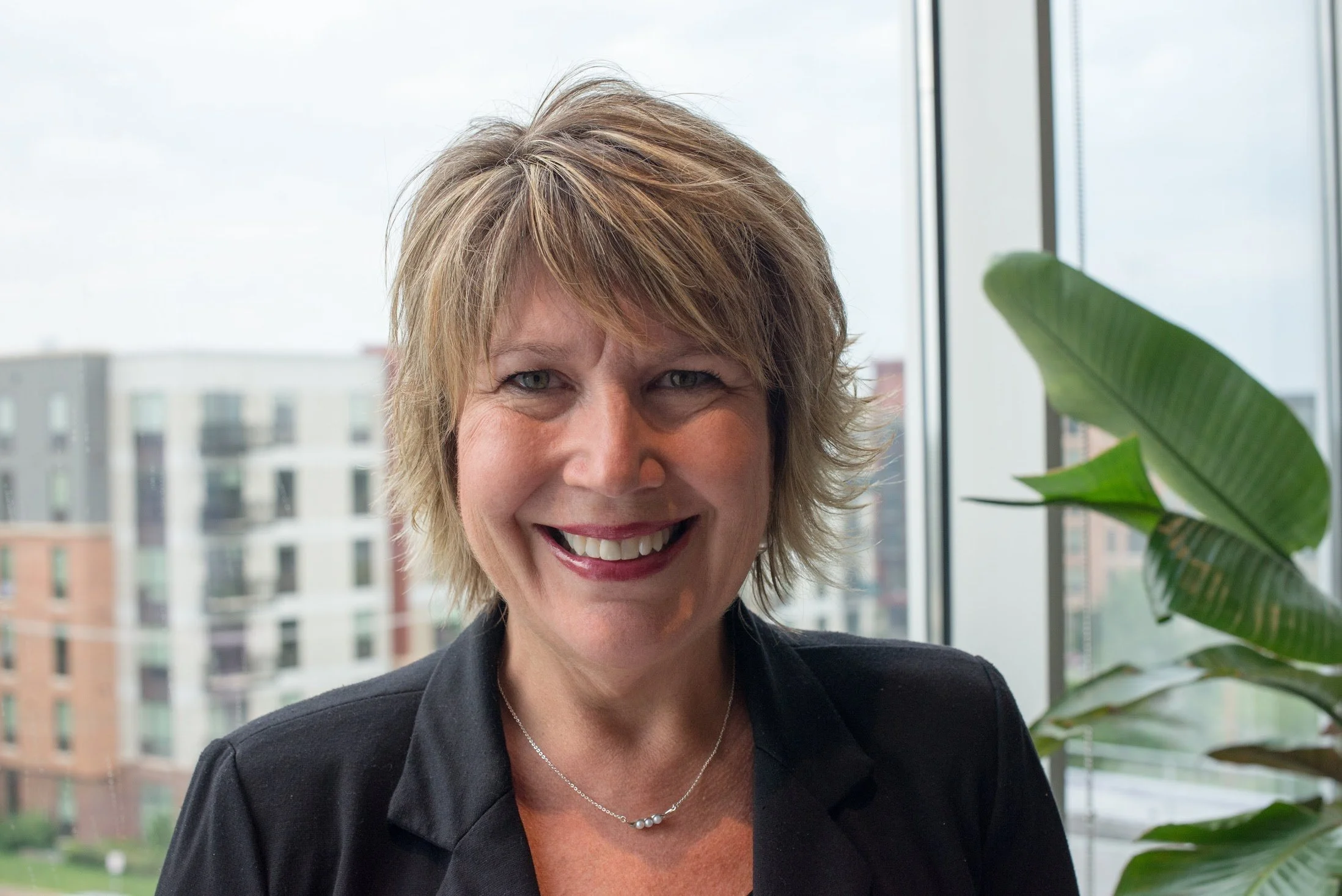 A smiling woman with short blond hair wearing a black blazer and a silver necklace, standing indoors near a window with a cityscape and a large green plant in the background.