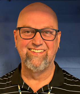 Close-up of a smiling man with a beard, glasses, and a bald head, wearing a black collared shirt with white stripes, against a blue background.