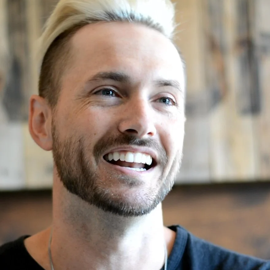 Smiling person with short light hair and beard in a casual setting.
