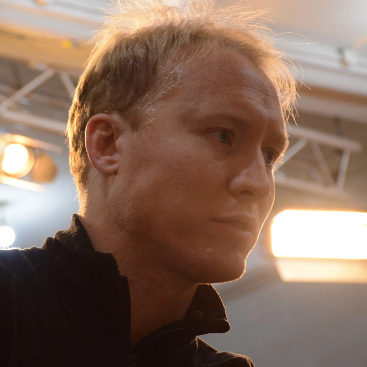 Close-up of Jay Denton with short blond hair, wearing a black jacket, with studio lights in the background.