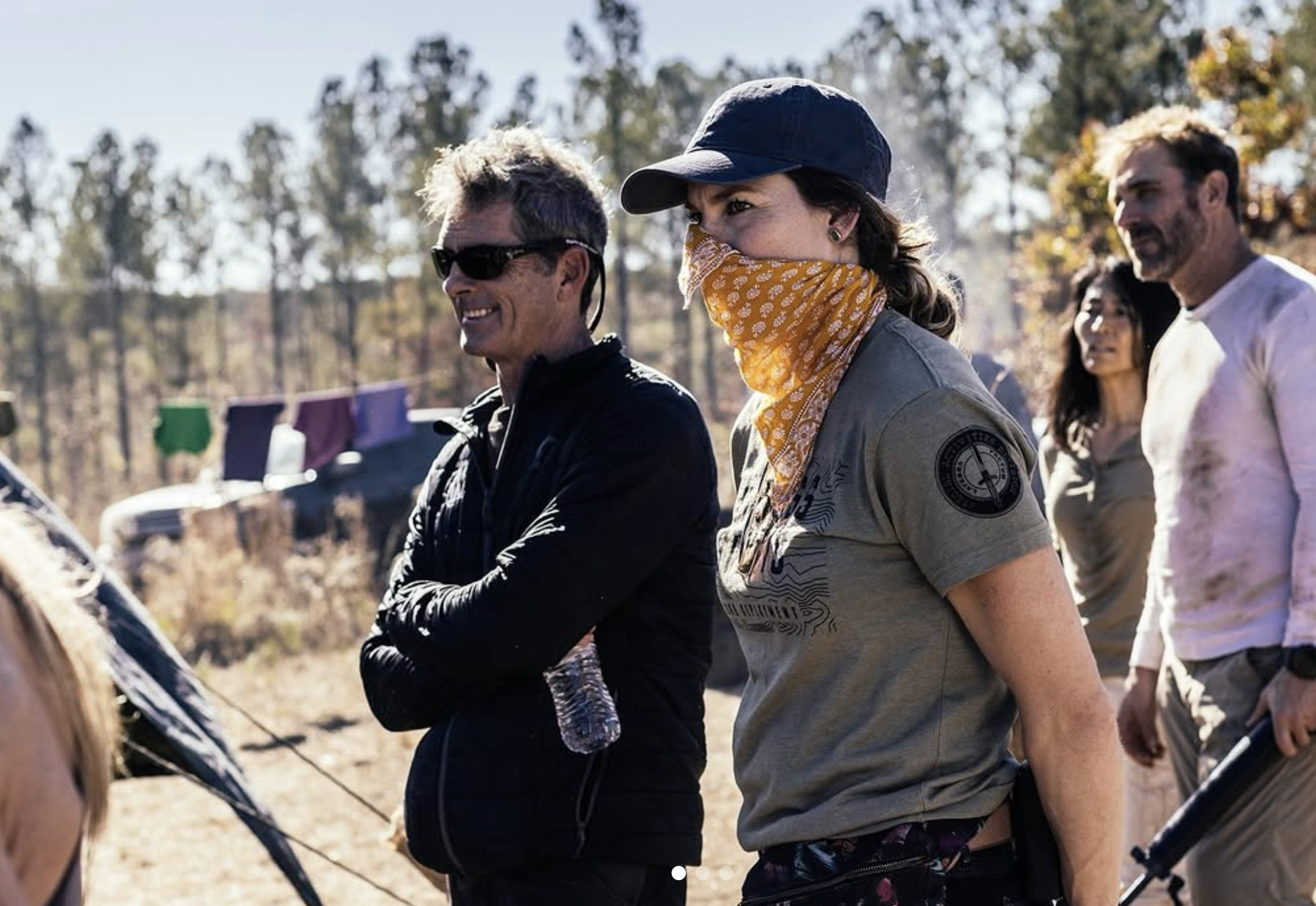 Group of people outdoors, some wearing casual and outdoor gear, one person with a bandana over their face. Trees in the background.