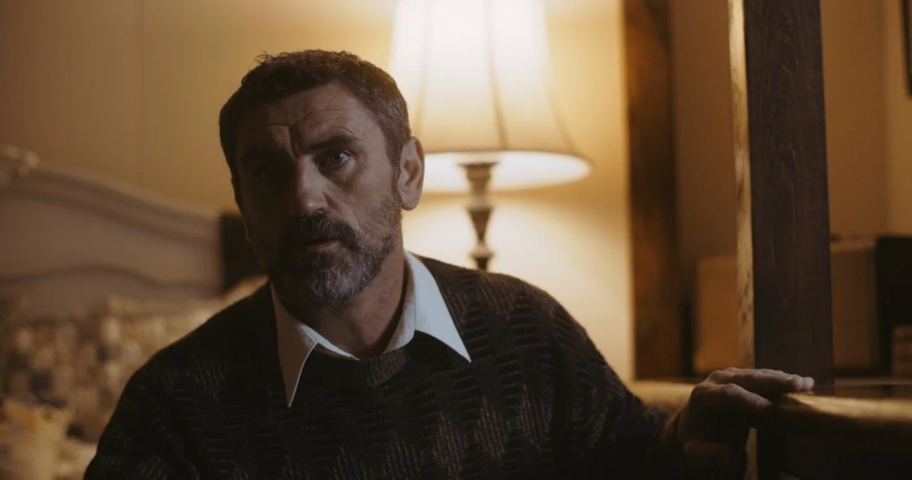 Man with a beard sitting indoors near a lamp