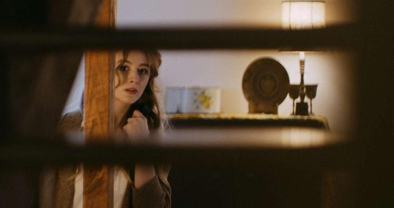 Woman peeking around a doorframe, with a softly lit room in the background featuring a lamp and decorative items on a table.