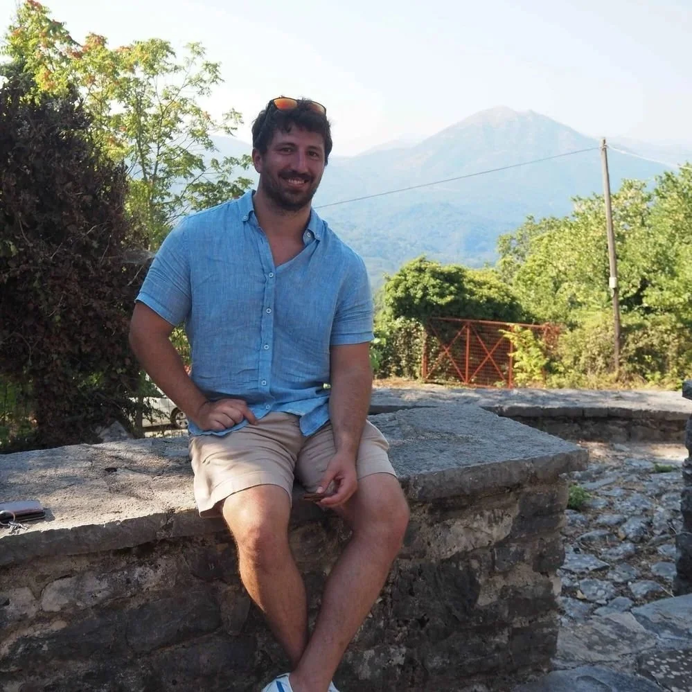 A man sitting on a stone ledge outdoors, wearing a blue shirt and tan shorts, with mountains and greenery in the background.