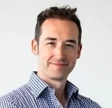Portrait of a man with short dark hair, wearing a checkered shirt, smiling slightly, against a plain white background.