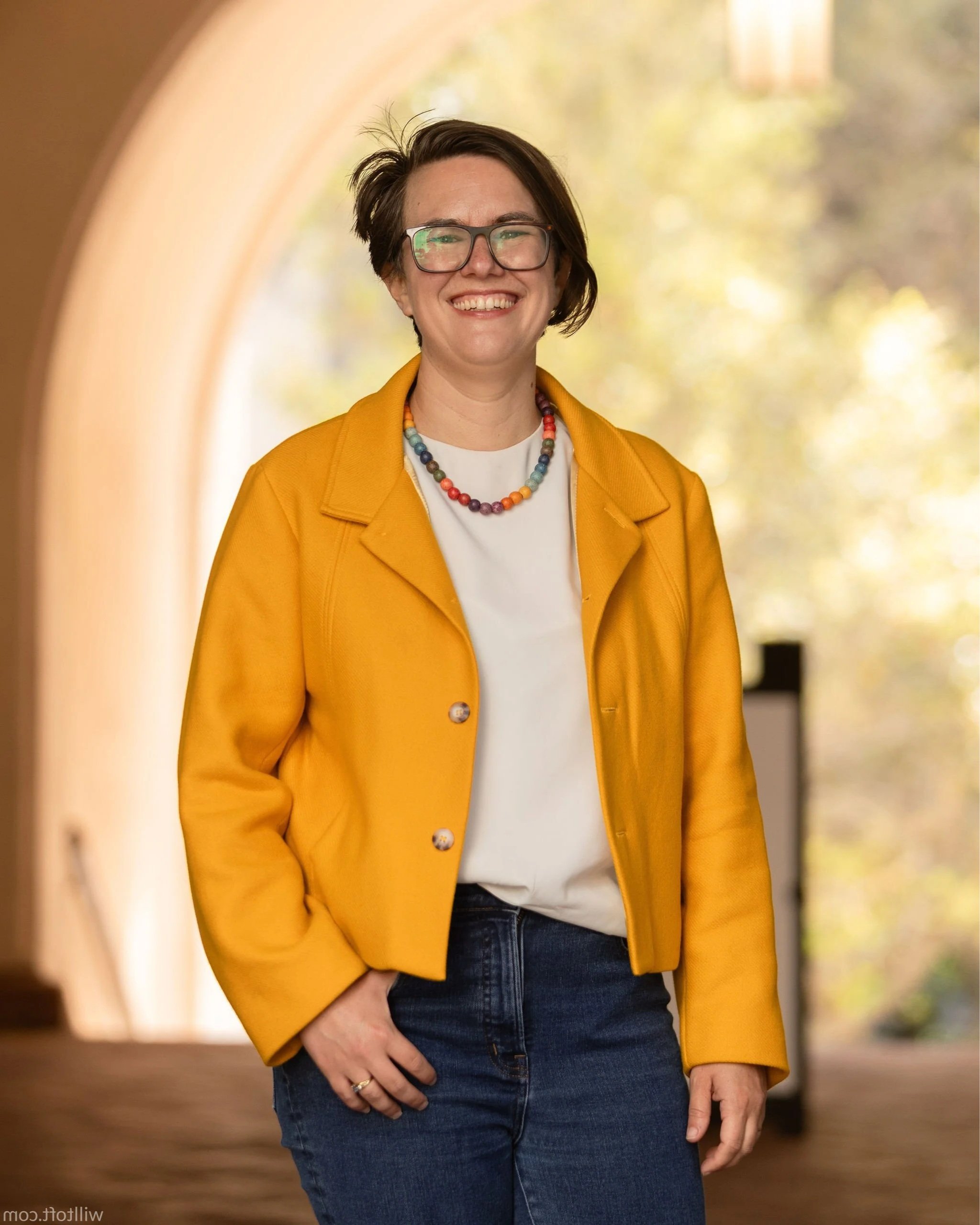 Close-up of a woman smiling, wearing glasses, a yellow jacket, a multicolored beaded necklace, a white inner shirt, and blue jeans, standing in an indoor corridor with a blurred background of warm indoor colors and autumn trees outside.
