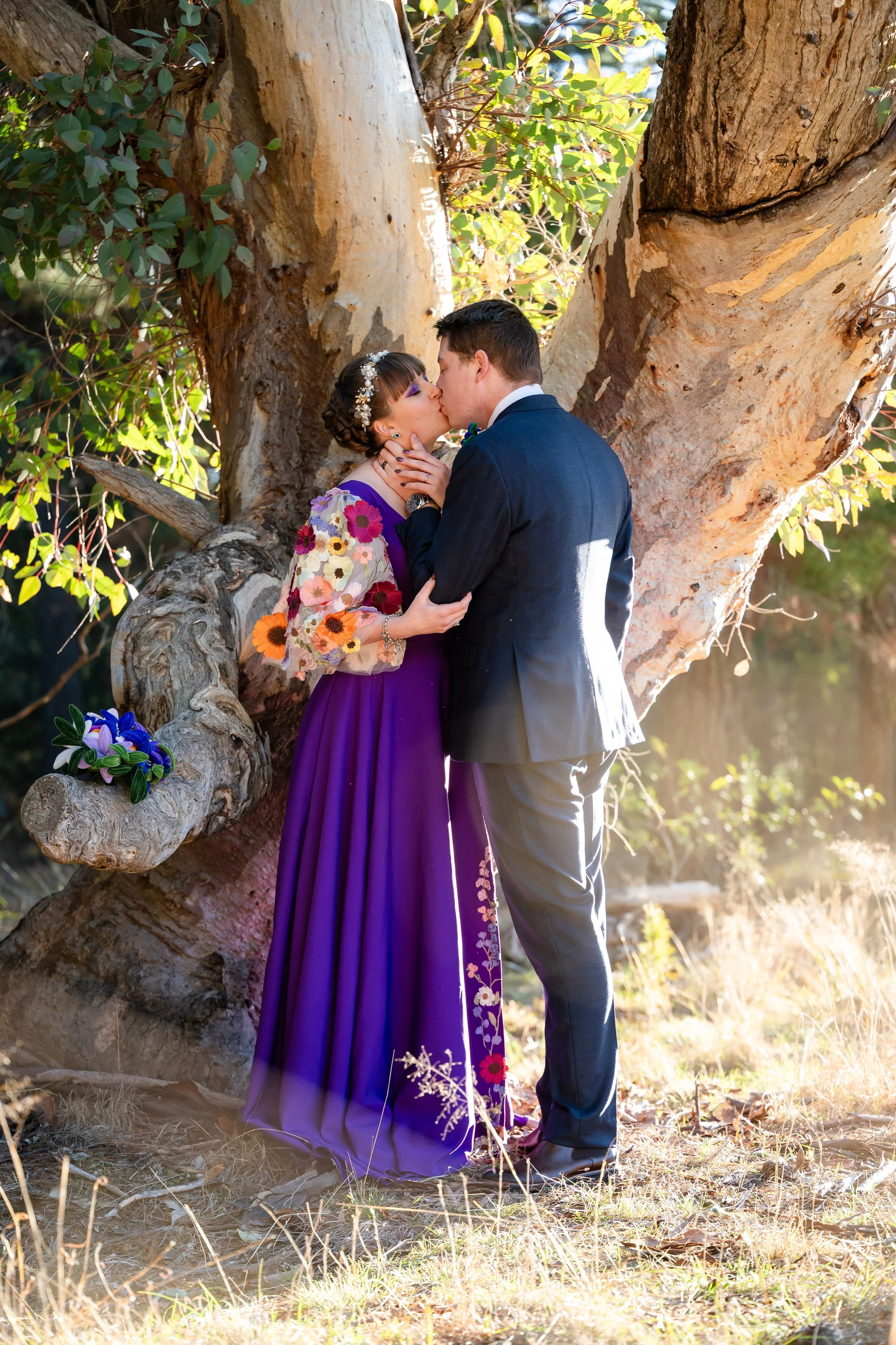 A couple kissing in front of a large tree, with the woman wearing a purple dress with floral embroidery and the man in a suit, during a sunny outdoor scene.