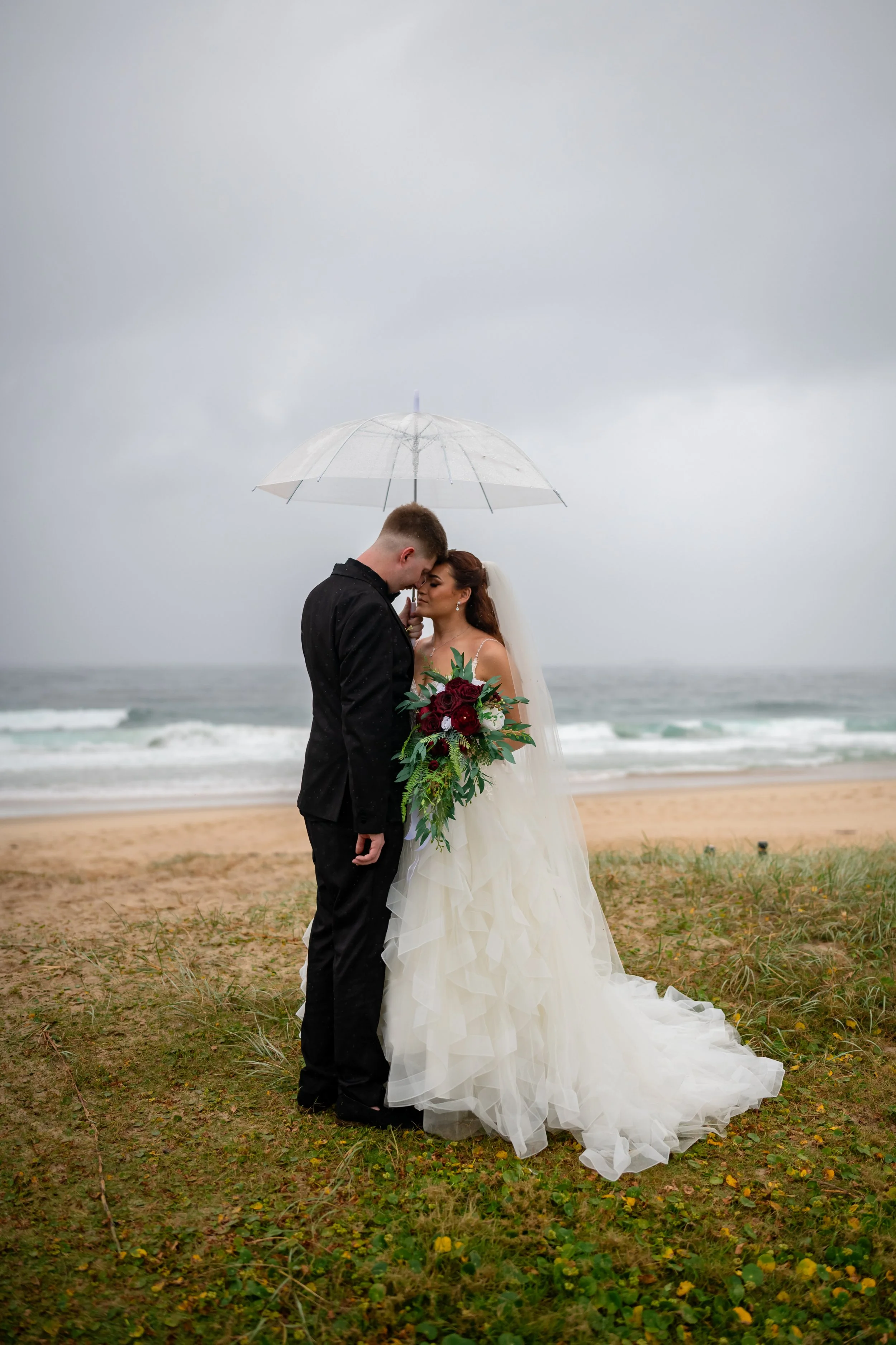 A bride and groom standing close together under a white umbrella on a rainy beach, sharing a romantic moment. The bride holds a large bouquet of red and white flowers with green foliage, and she wears a white wedding gown with a veil. The groom is in