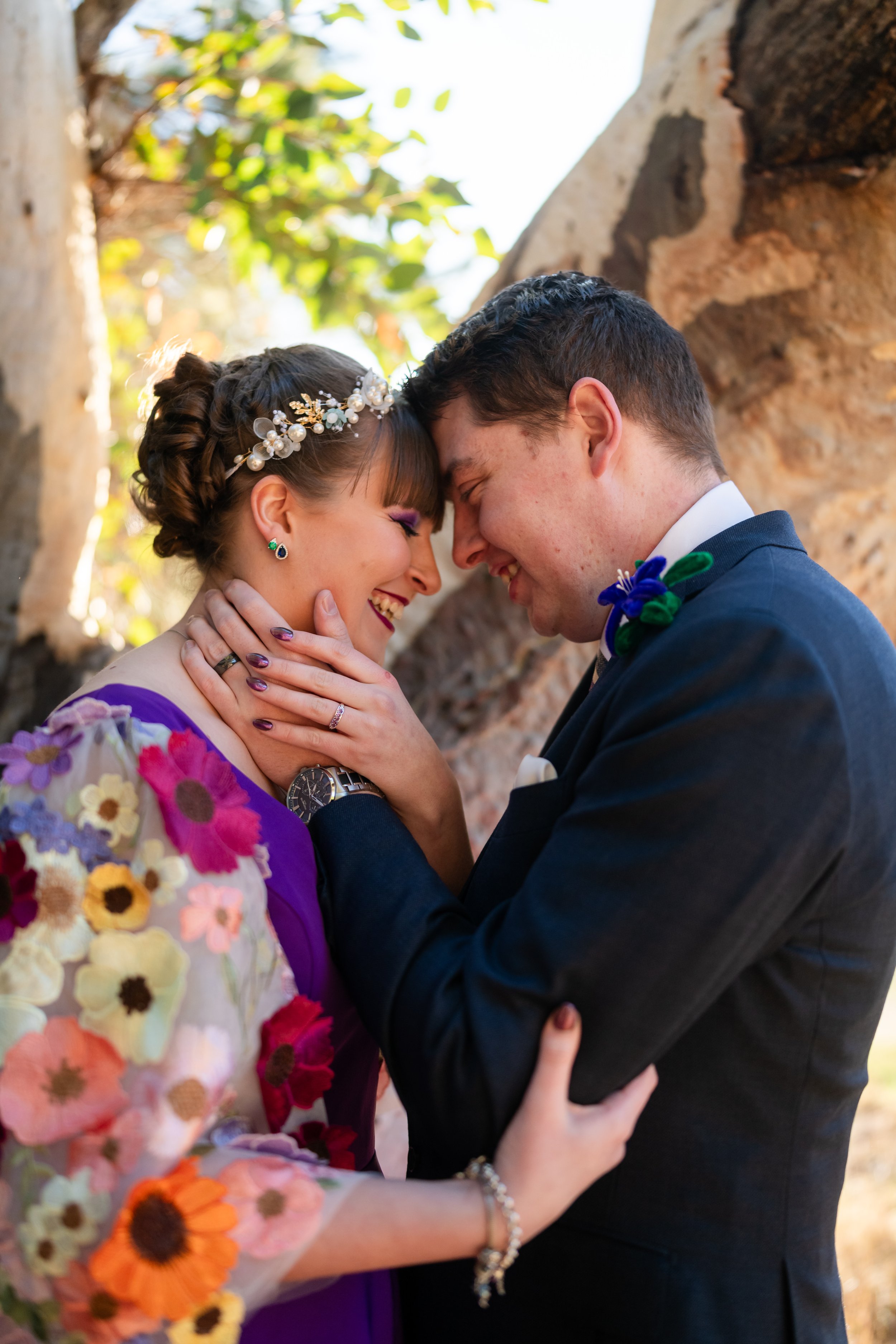 A newlywed couple sharing an intimate moment outdoors, with their foreheads touching and closed eyes, smiling happily against a background of trees and rocks.