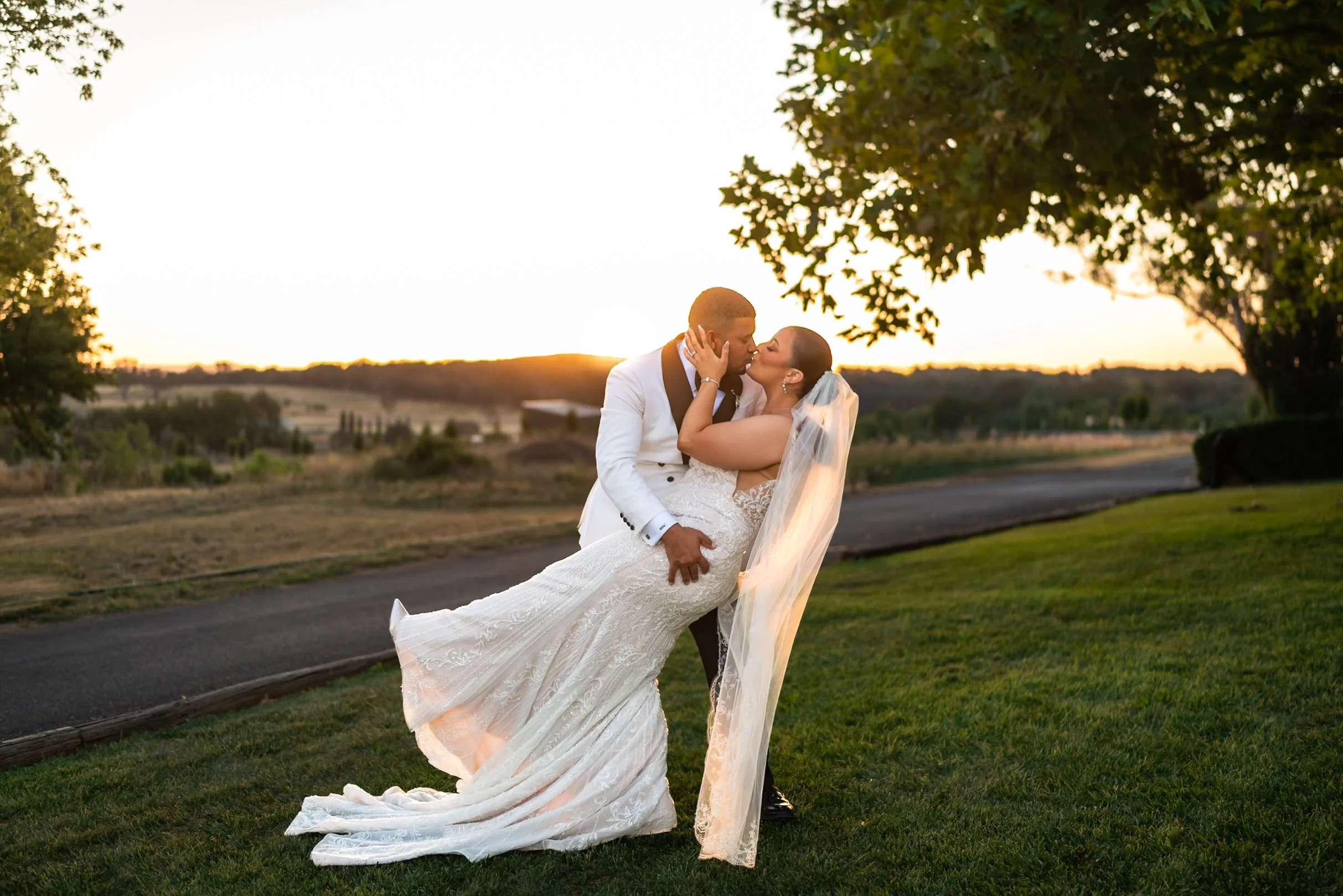 Groom in a white tuxedo dips his bride for a kiss at sunset on the lawn at Shaw Estate, Murrumbateman, her veil and train spread across the grass