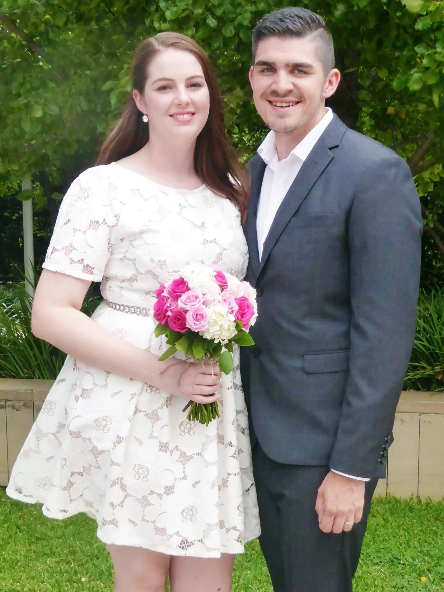 A woman in a white lace dress holding a bouquet of pink and white roses, standing next to a man in a gray suit and white shirt, outdoors with green foliage in the background.
