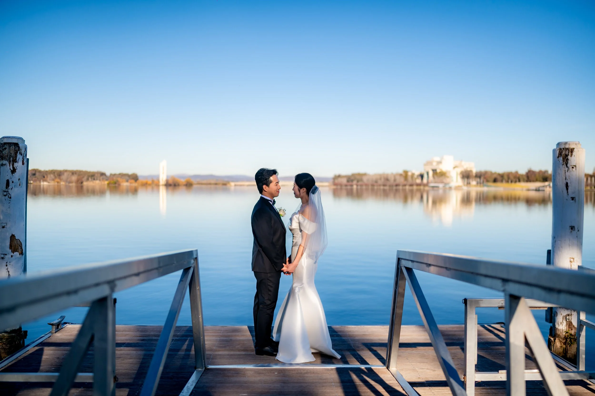 A bride and groom stand facing each other on a wooden dock by a calm lake, holding hands, with a clear blue sky overhead.