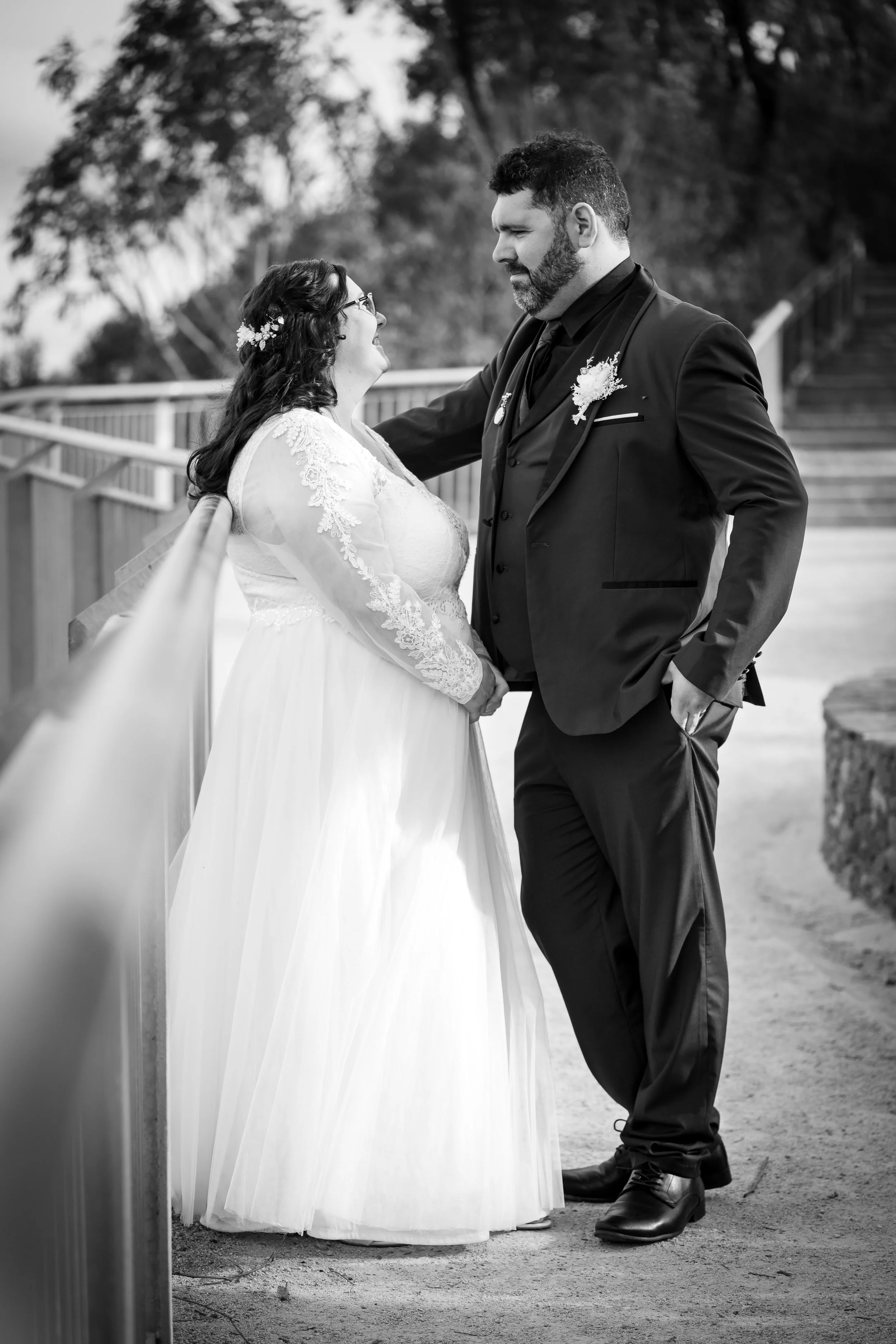 Black and white photo of a bride and groom standing outdoors, gazing at each other. The bride wears a lace long-sleeve wedding dress, and the groom wears a dark suit with a boutonniere. The setting includes a railing and trees in the background.