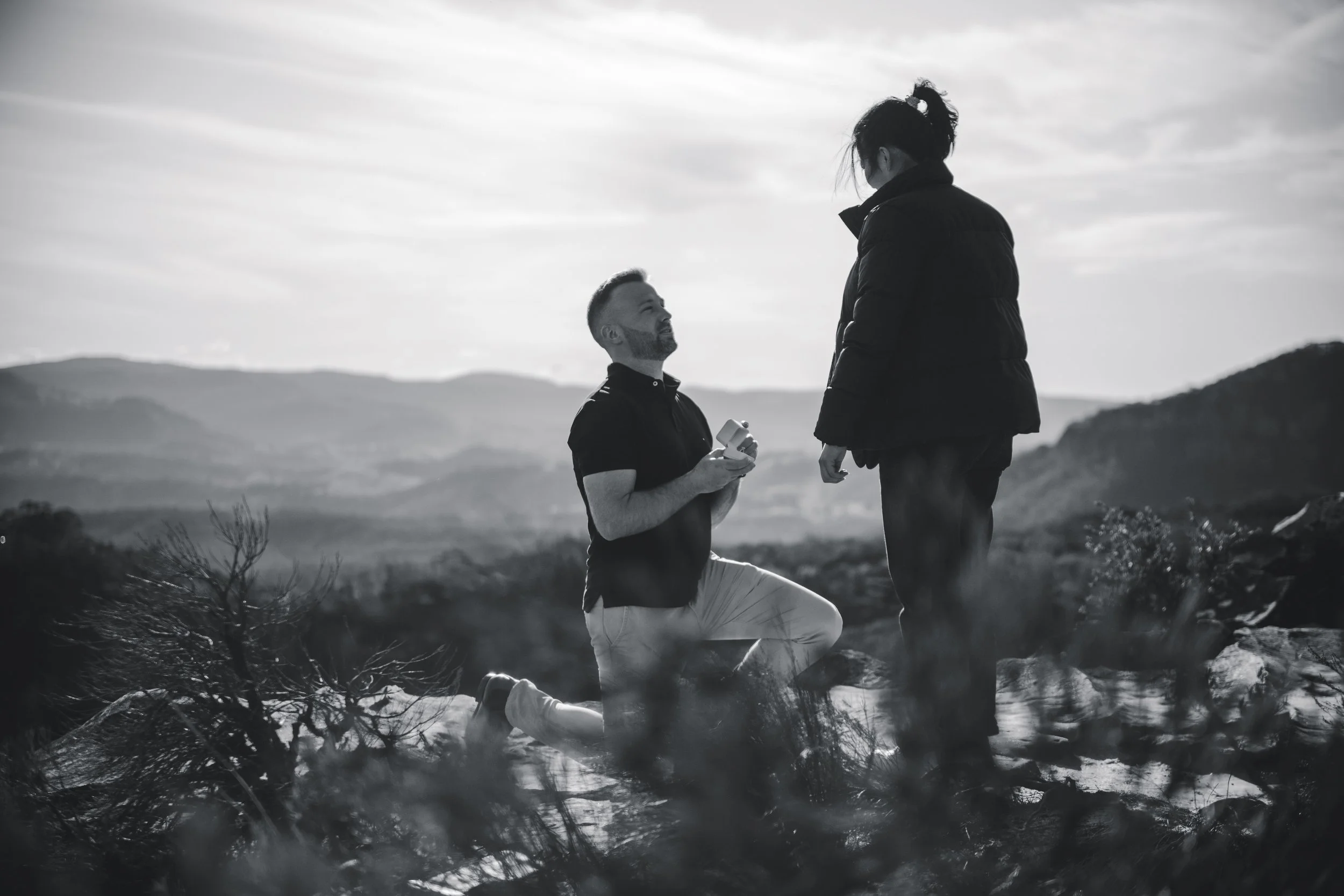 A man kneeling on one knee proposing to a woman outdoors with mountains in the background, in black and white.