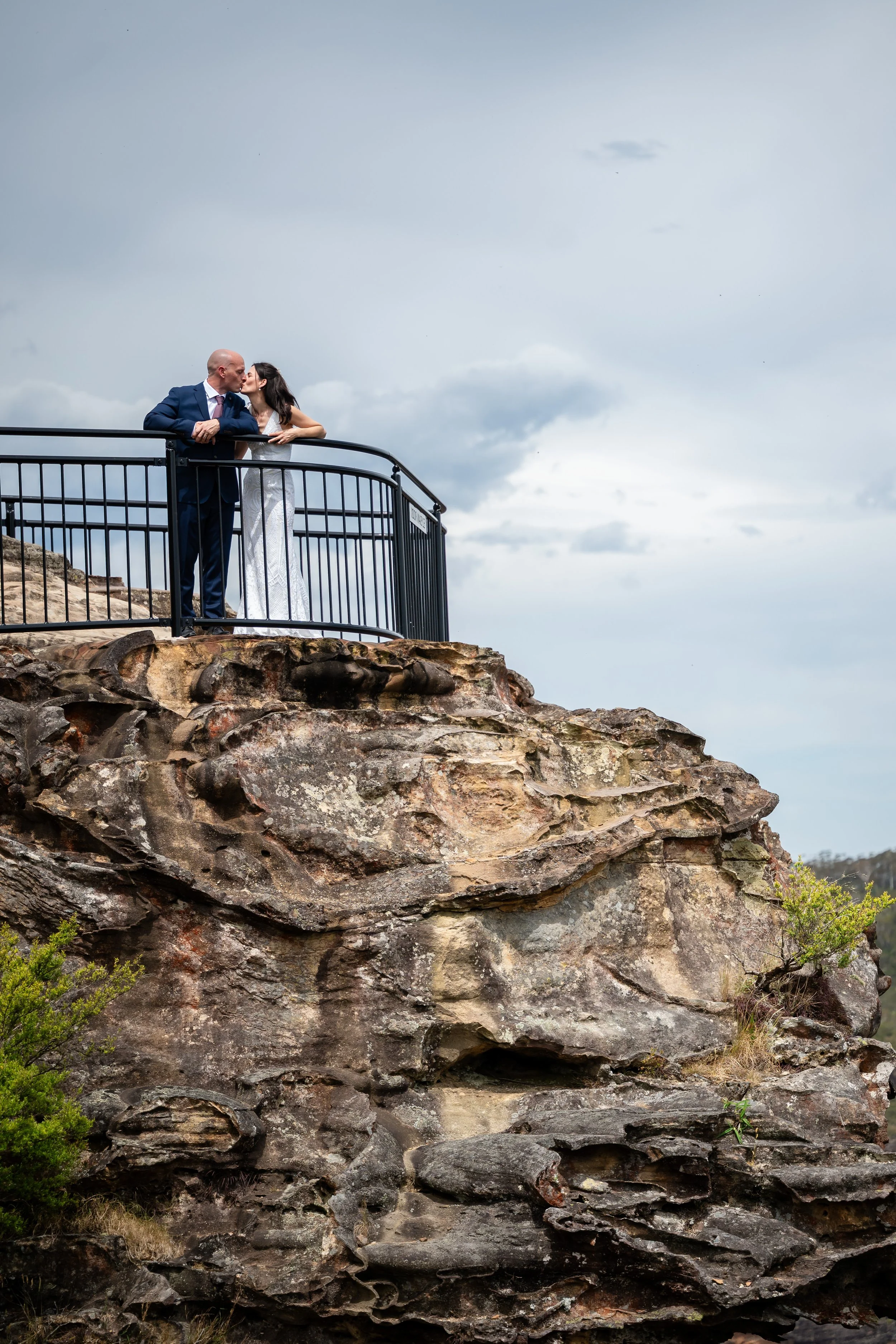 A couple dressed in formal attire sharing a kiss on a viewing platform on a rocky cliffside.