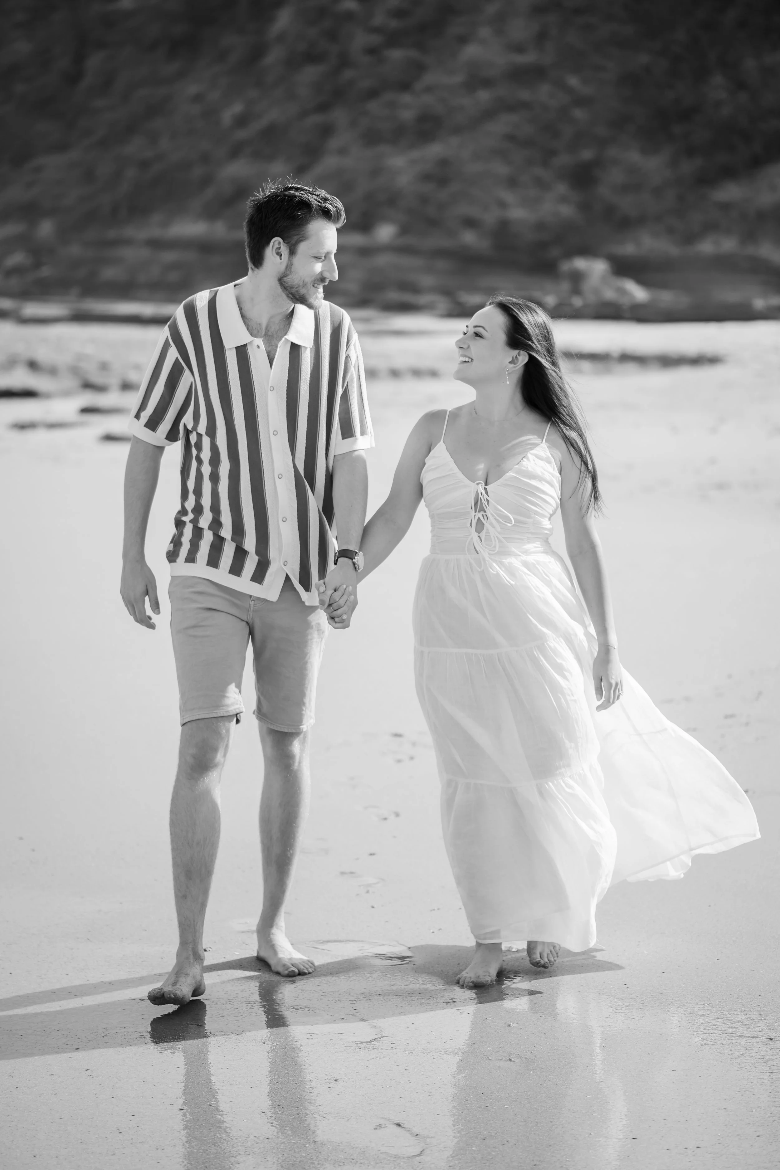 A couple walking hand-in-hand on the beach, smiling and looking at each other, with mountains and water in the background.